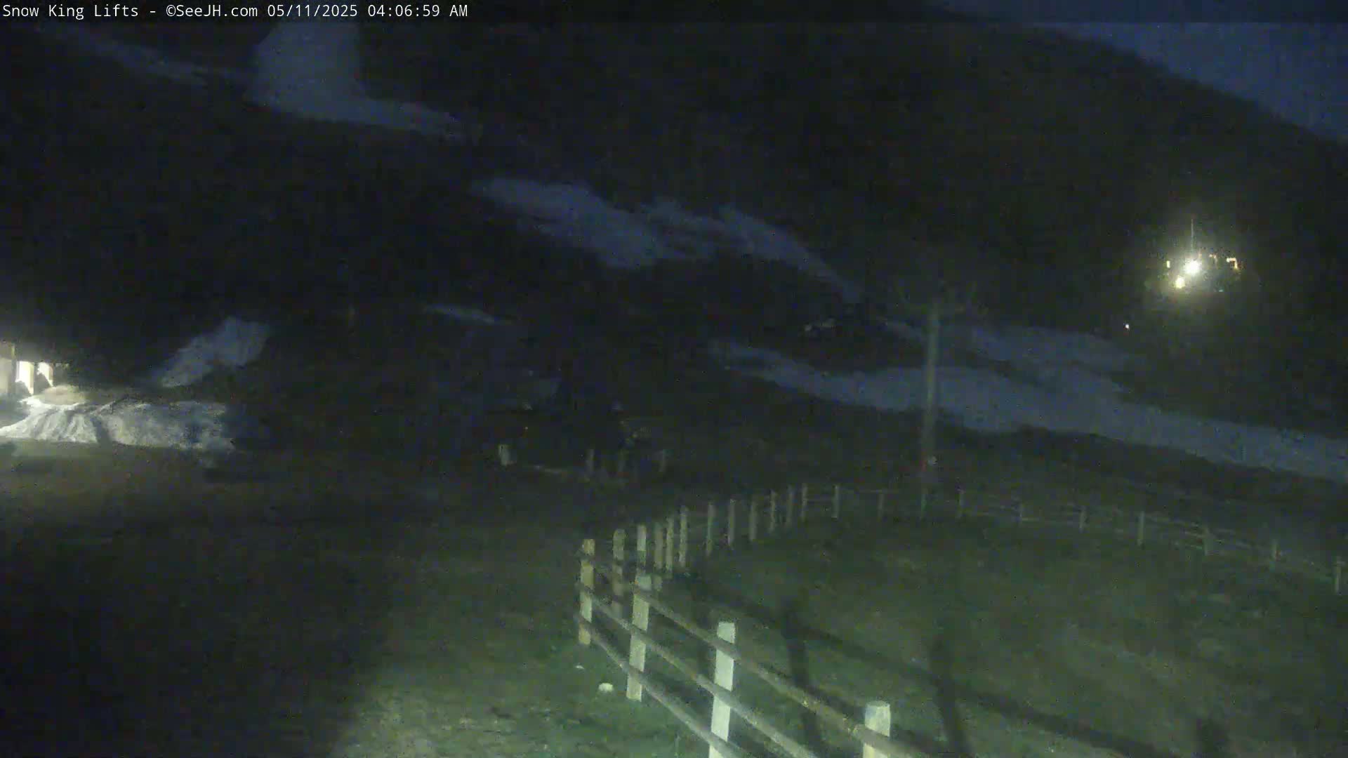 A nighttime, low-light view of a snow-dusted hill and a wooden fence, with a small light source visible in the distance.