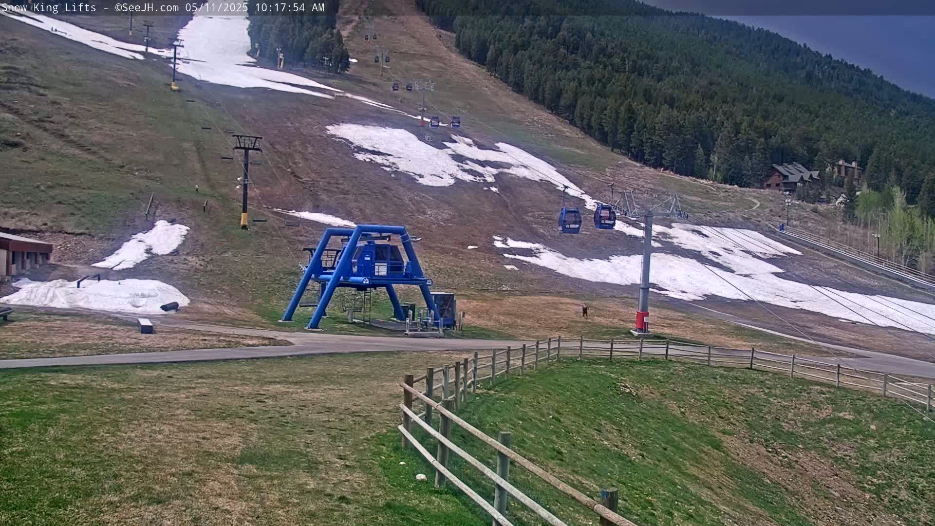 A mostly bare ski slope with patches of snow, a blue ski lift terminal, and a chairlift carrying passengers is visible under partly cloudy skies.