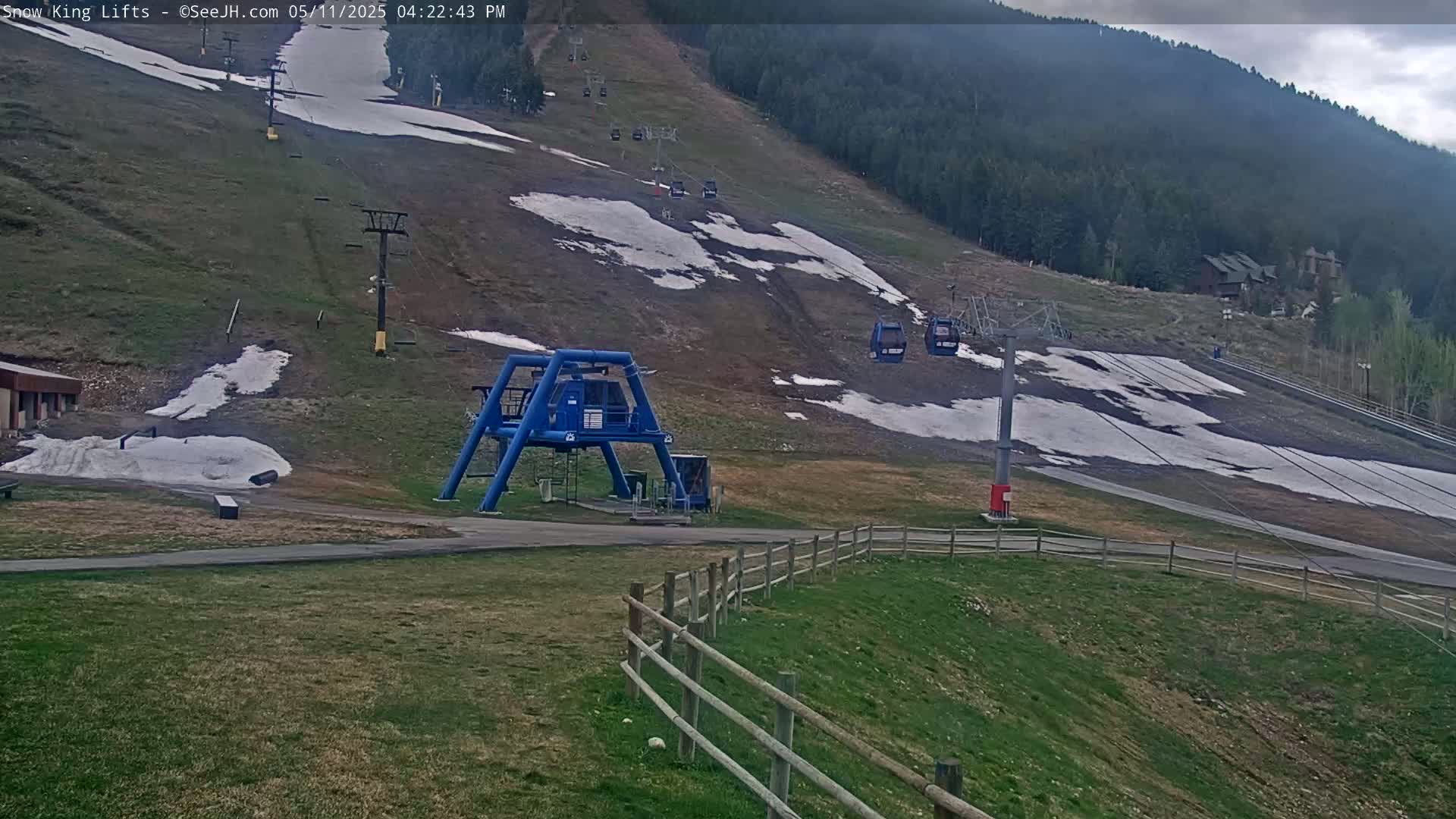 A ski lift base station and chairlifts ascend a grassy hill with patches of melting snow under an overcast sky.