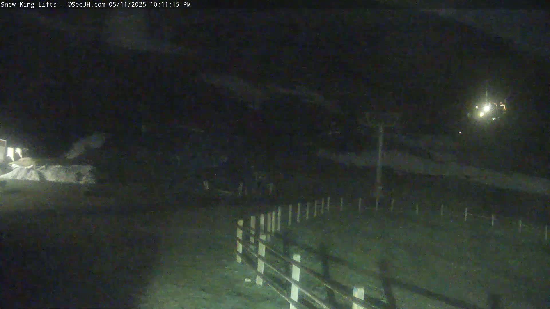A nighttime, low-light view of a snow-covered area with a wooden fence and a distant light source.