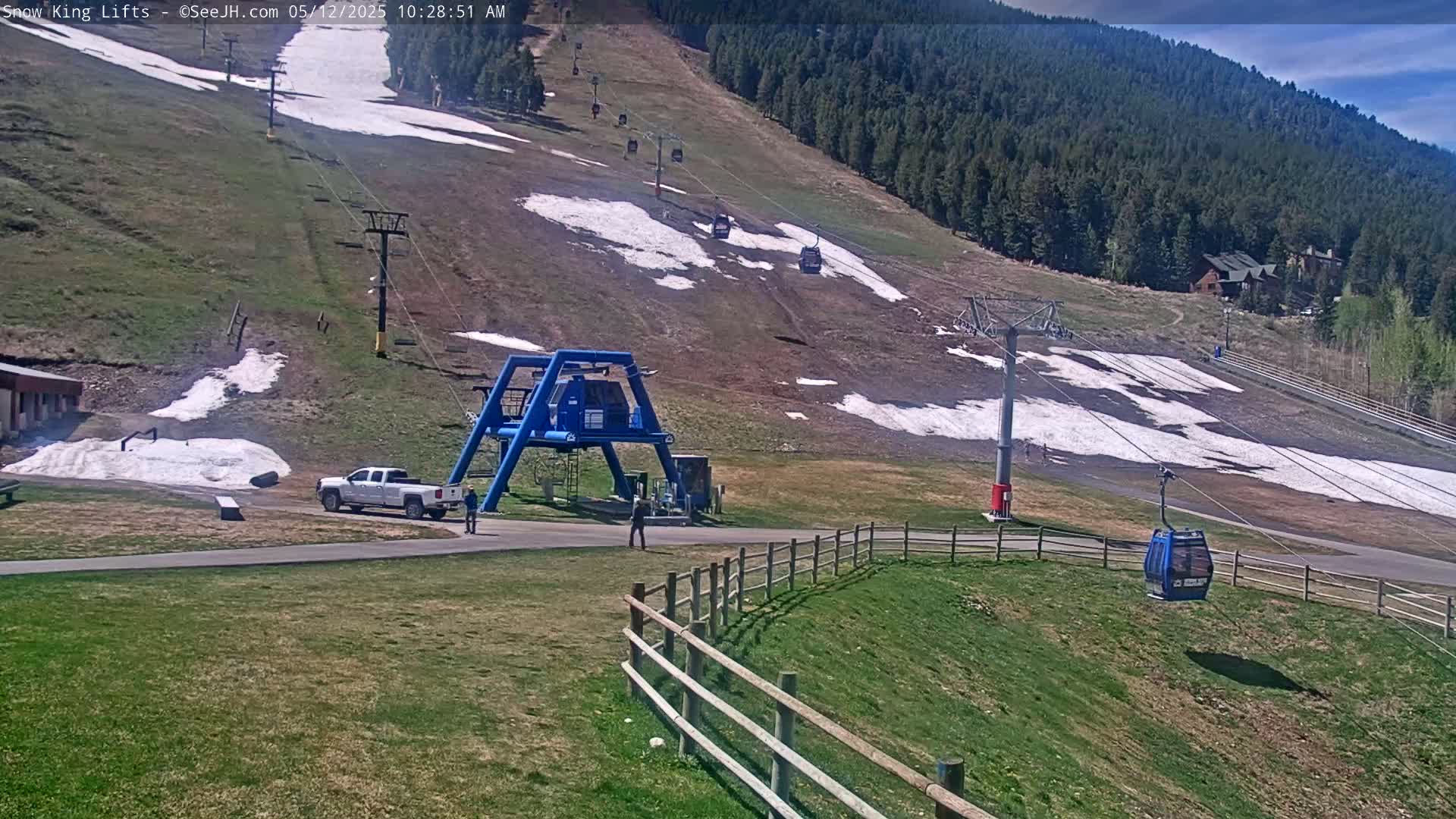 A ski lift with several empty blue gondolas ascends a grassy hill with patches of remaining snow under a partly sunny sky.