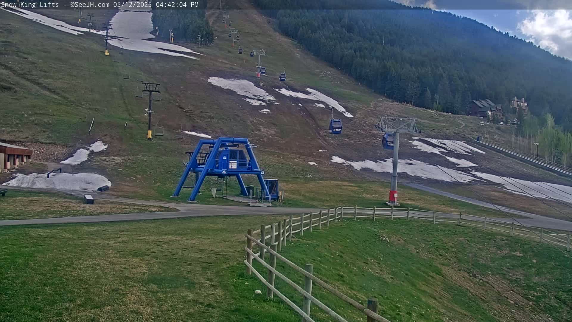 A mostly green ski slope with patches of melting snow, ski lifts, and a blue chairlift terminal under a partly cloudy sky.