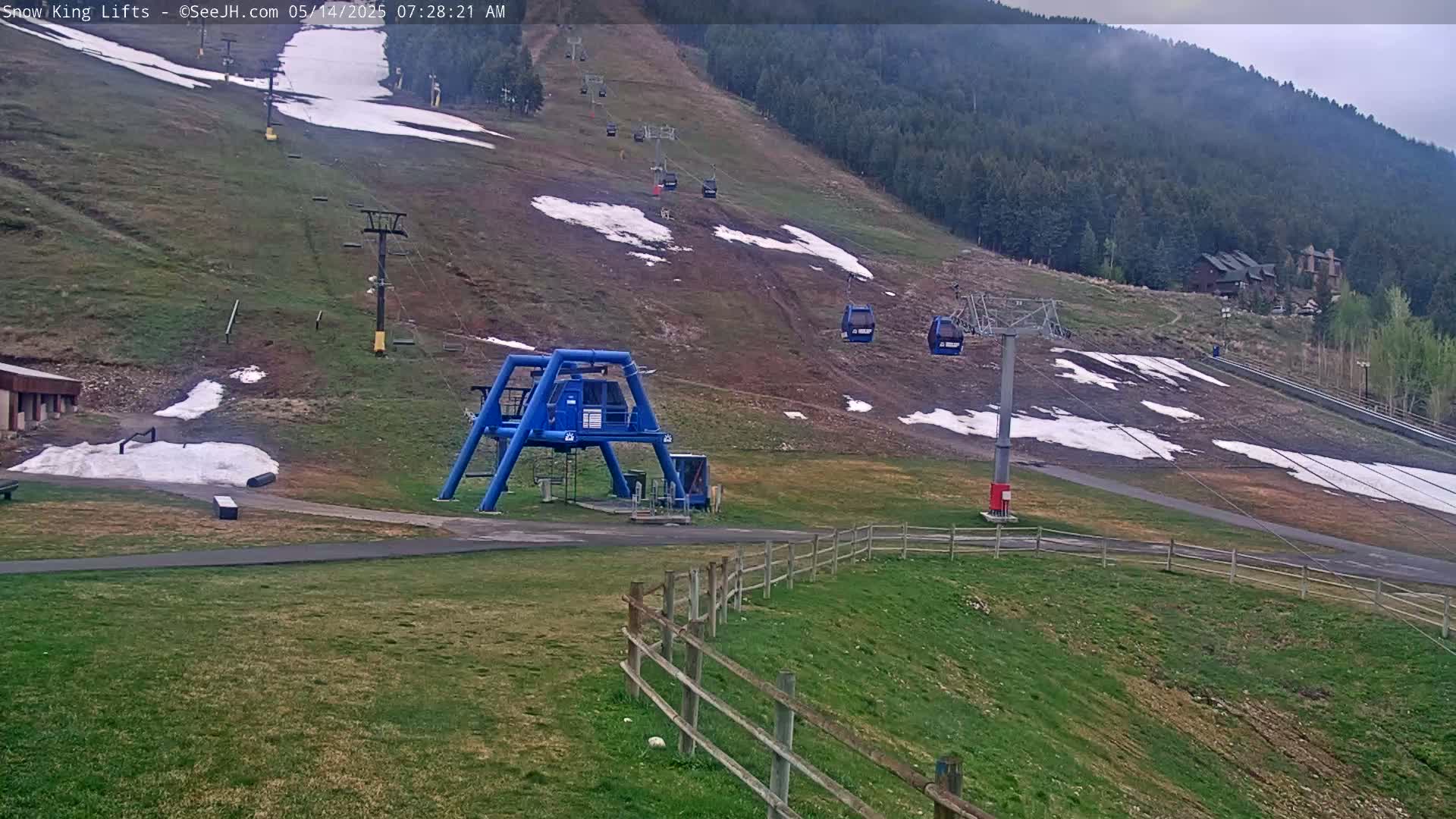 A ski lift base station with a blue structure sits in a grassy area next to a wooden fence, overlooking a mountainside with patches of melting snow and several chairlifts ascending the hill on an overcast day.