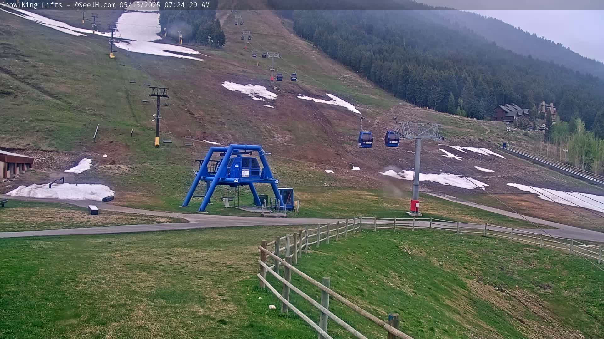A ski lift base station and chairlifts ascend a grassy mountainside with patches of snow under an overcast sky.
