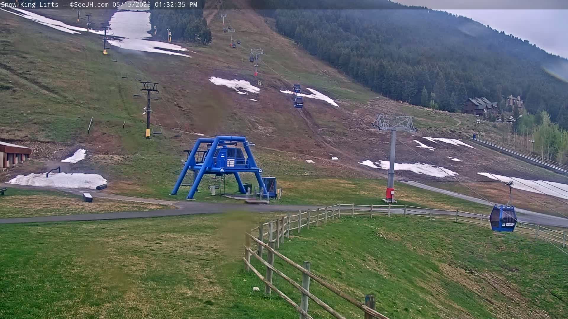 A ski lift and chairlift ascend a grassy hillside with patches of remaining snow under an overcast sky.