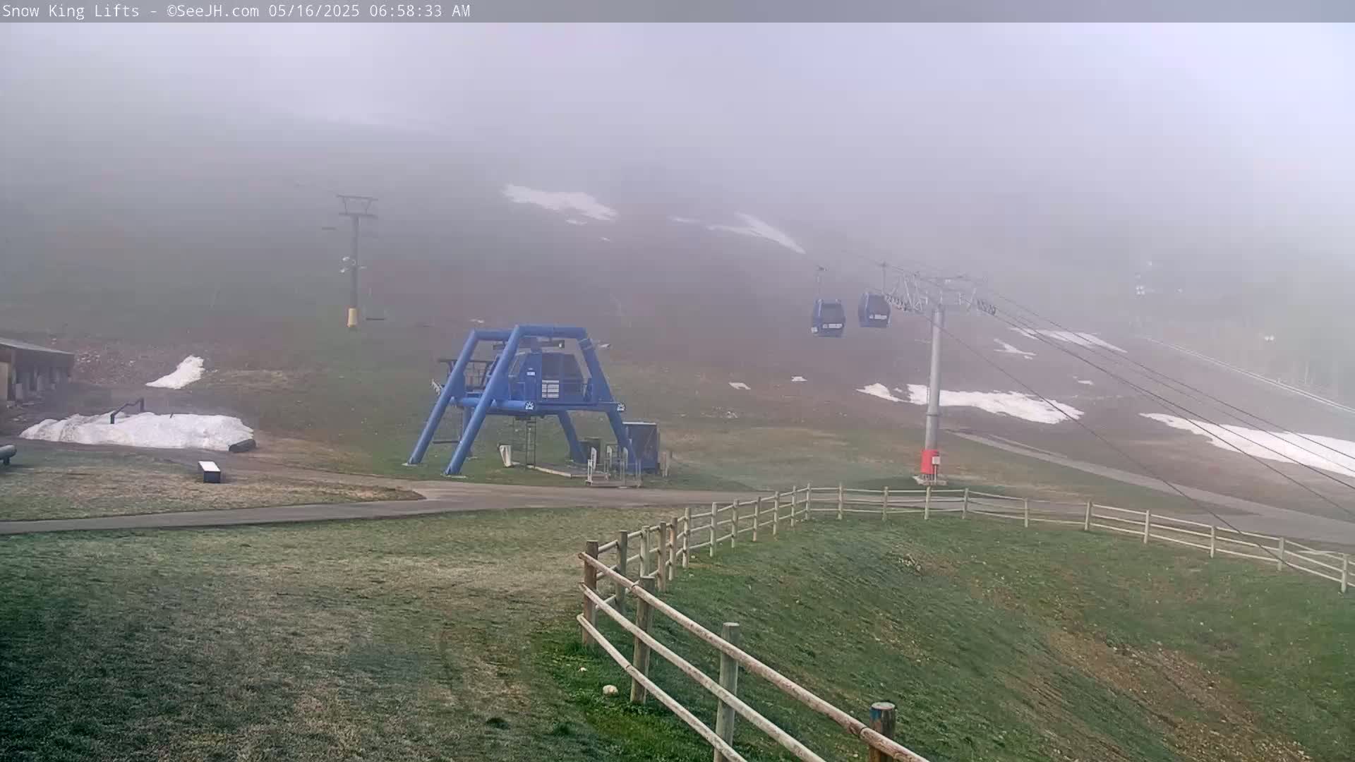 A foggy scene shows a ski lift with a few passenger cars, a ski lift base station, and patches of snow on a grassy hill.