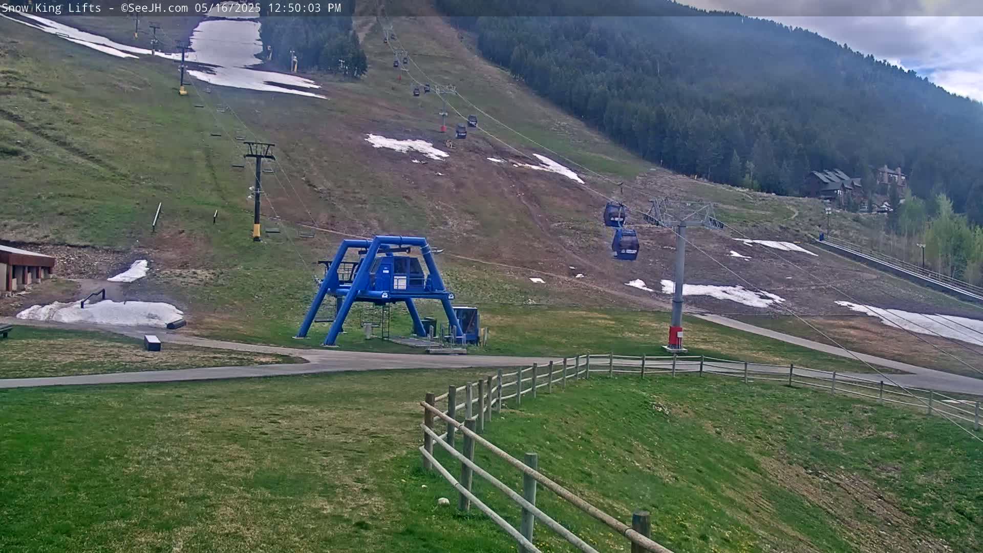 A ski lift with several empty gondolas ascends a grassy mountainside with patches of remaining snow under partly cloudy skies.