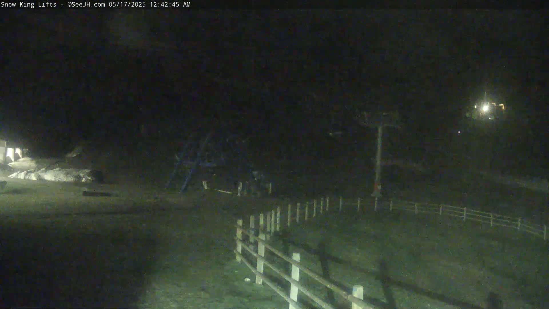 A nighttime, low-light view shows a ski lift and a snow-covered area, with a wooden fence in the foreground.