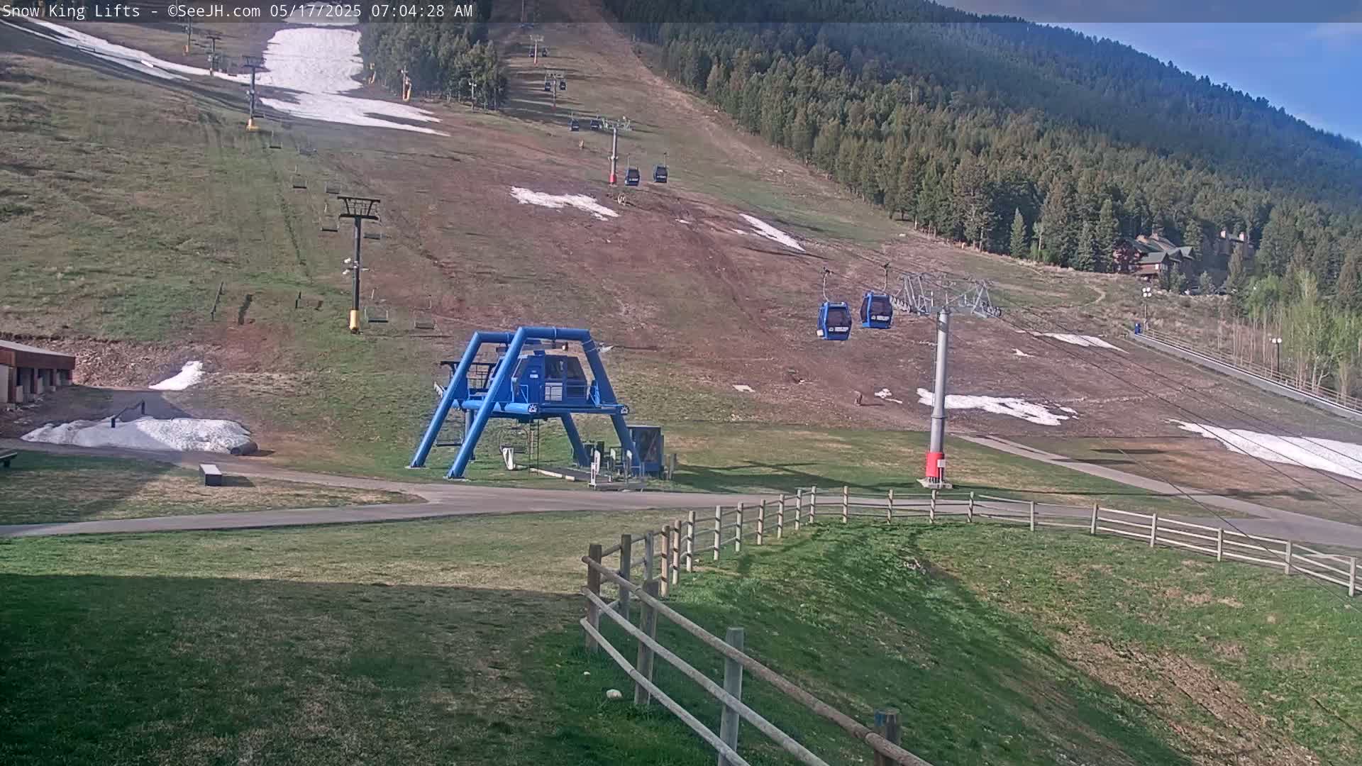 A ski lift base station and chairlifts are visible on a mostly brown grassy hill with patches of snow under a mostly clear sky.