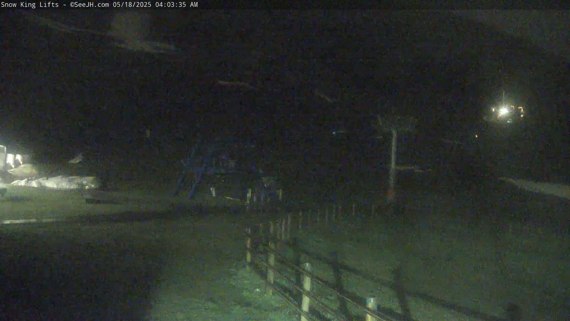 A nighttime, low-light view of a snow-covered area with a fence and some structures visible in the distance.