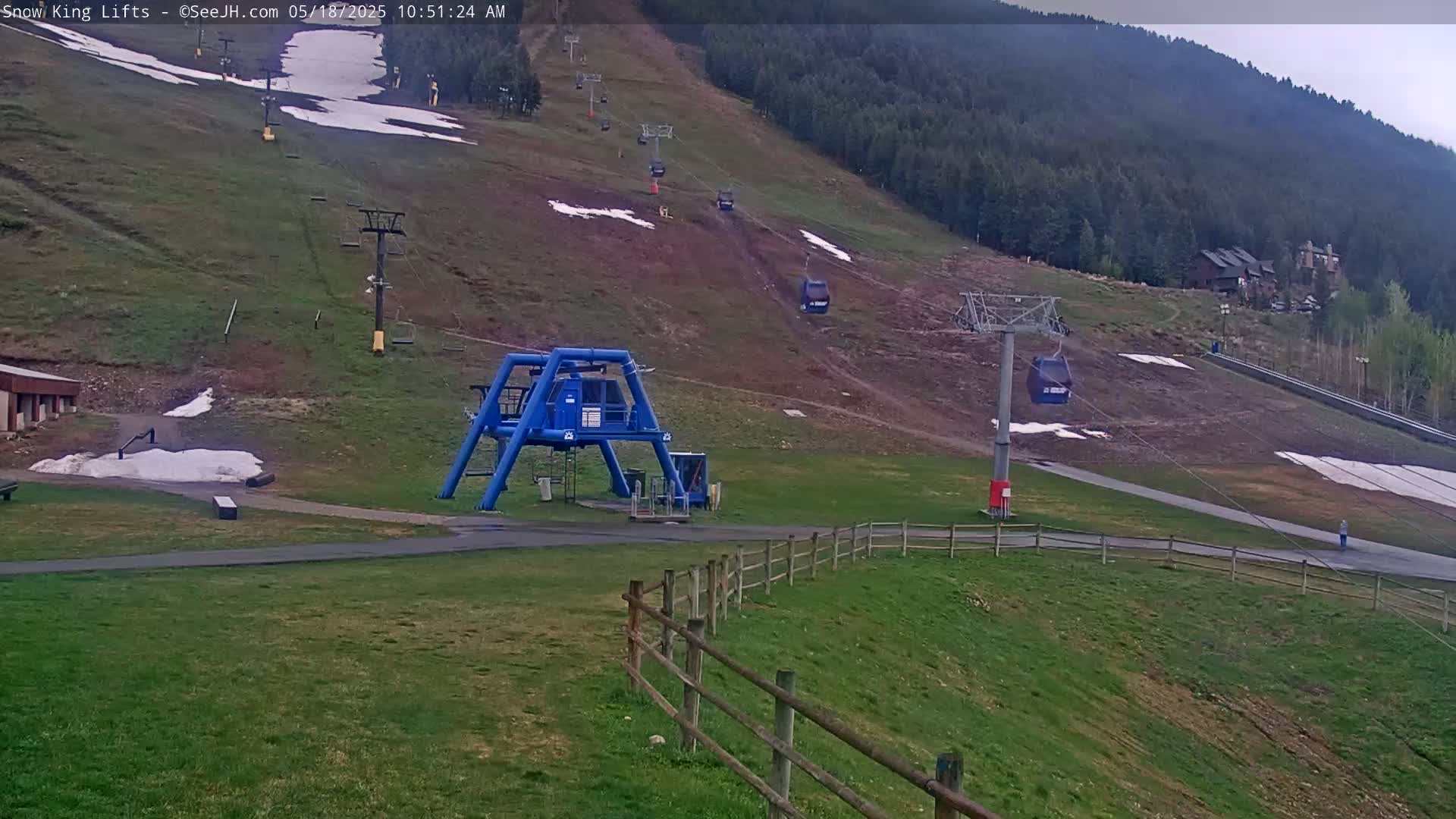A ski lift base station with several chairlifts ascending a grassy hill dotted with patches of melting snow, under an overcast sky.