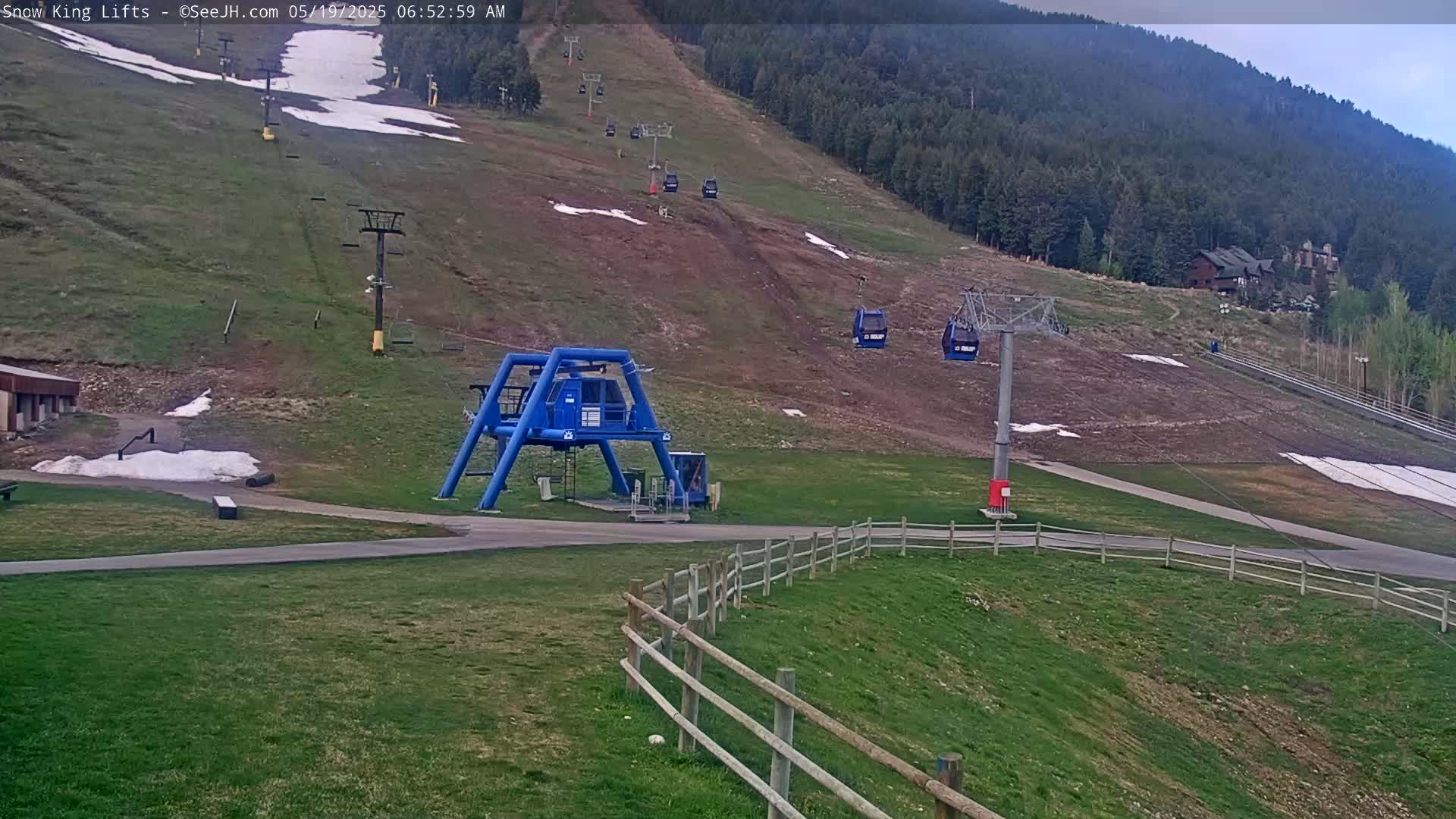 A ski lift base area with a mostly clear sky,  shows patchy snow on the grassy hill, with several chairlifts ascending the mountainside.