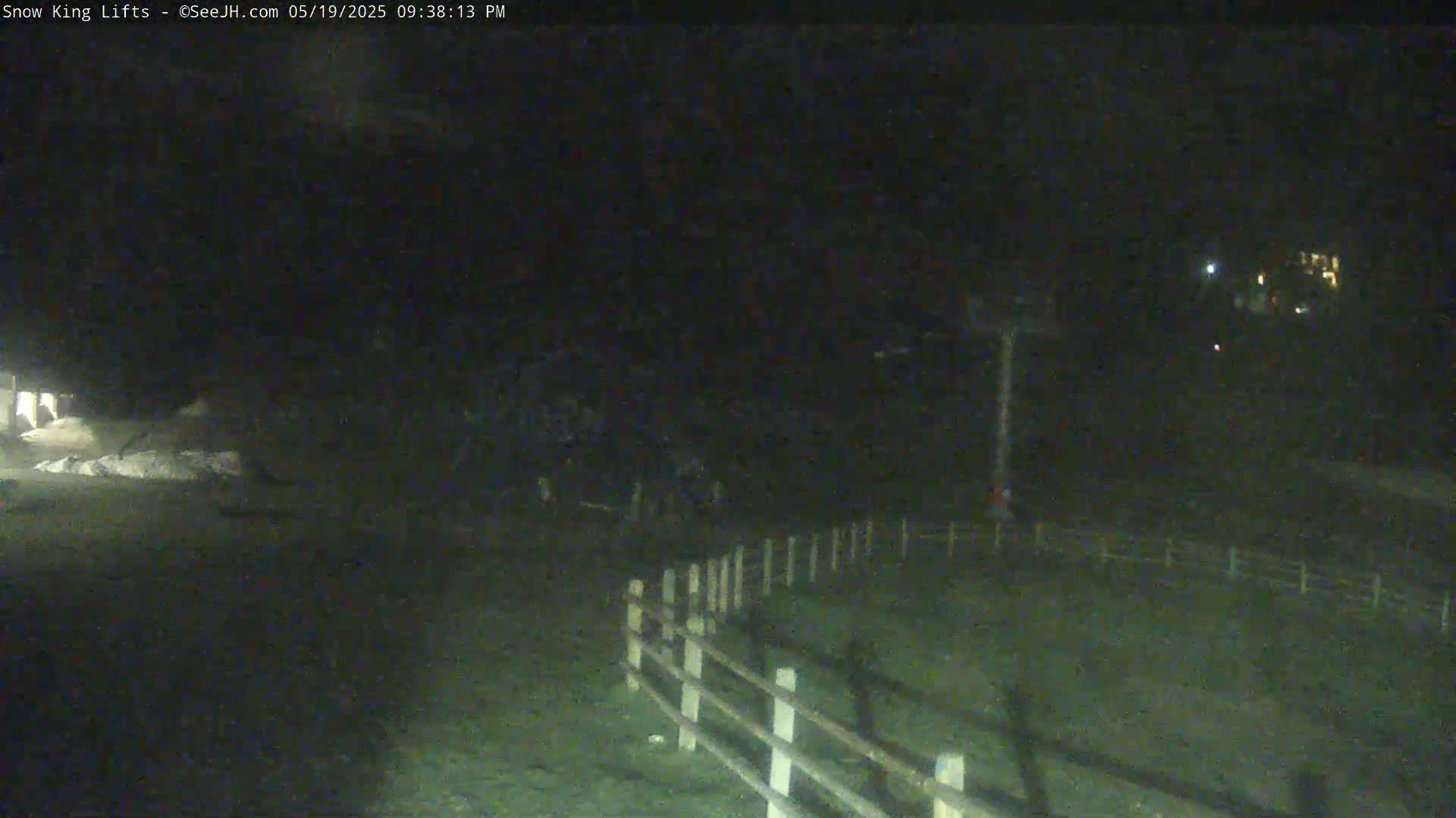 A nighttime, low-light view shows a section of a wooden fence next to a dark, grassy area, with a tall structure visible in the midground and faint lights in the far background.