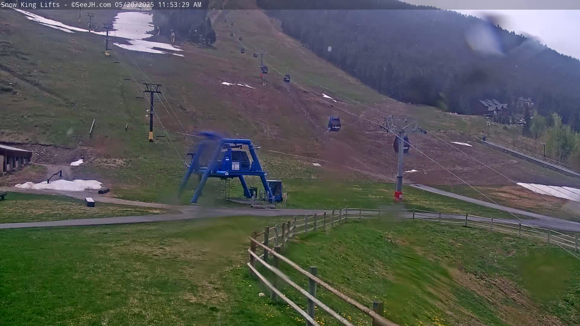 A ski lift with several empty chairlifts visible during rainy weather, set against a grassy hillside with patches of remaining snow.