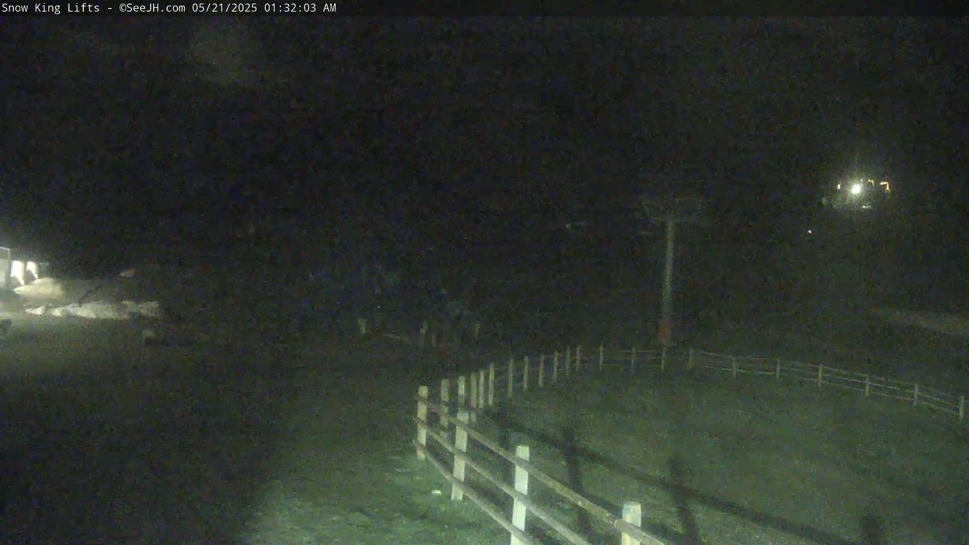 A nighttime, low-light view shows a section of a wooden fence running diagonally across the bottom of a dark, grassy area, with a ski lift in the background under a dark sky.