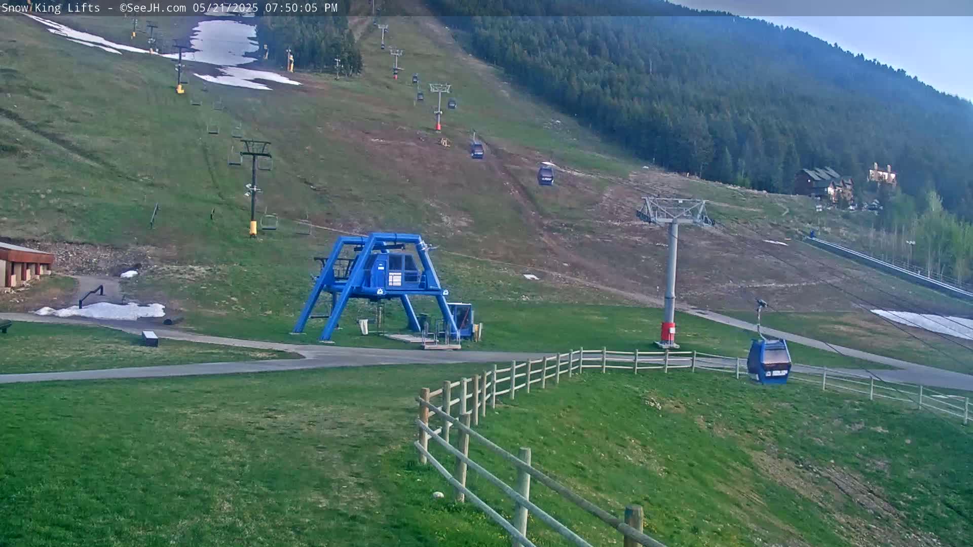 A ski lift base station and several empty chairlifts are situated on a grassy hillside under a partly cloudy sky, with remnants of snow visible in patches.