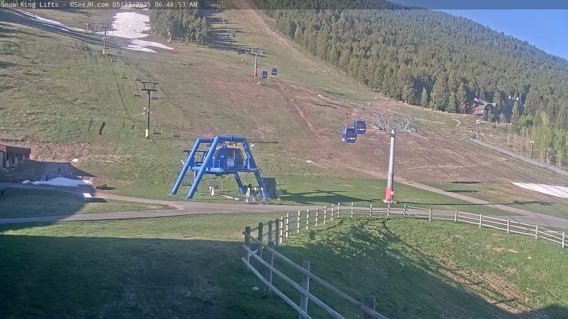A mostly sunny view of a ski lift area with chairlifts and a base station,  brown grass,  and some patches of remaining snow.