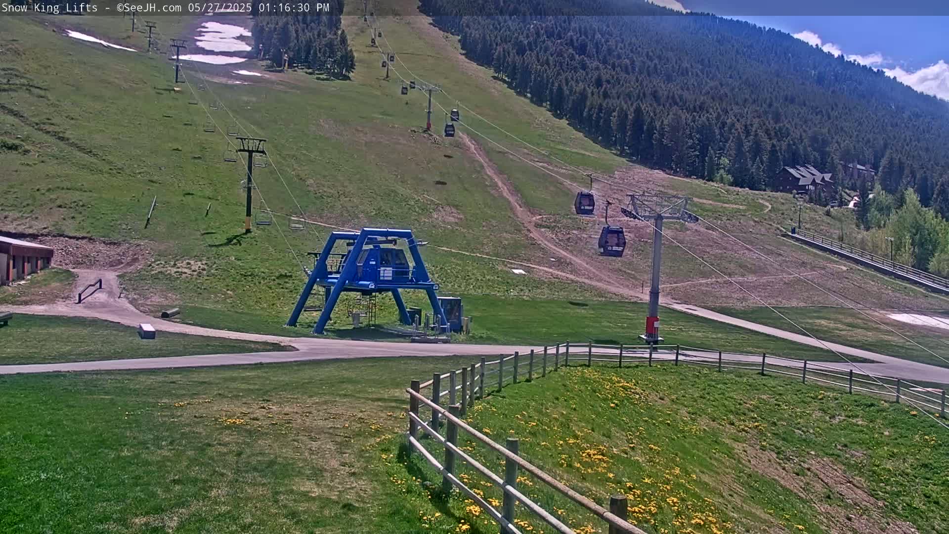 A ski lift base station and chairlifts are visible on a grassy hillside under a sunny sky, with a few patches of remaining snow visible higher up the mountain.