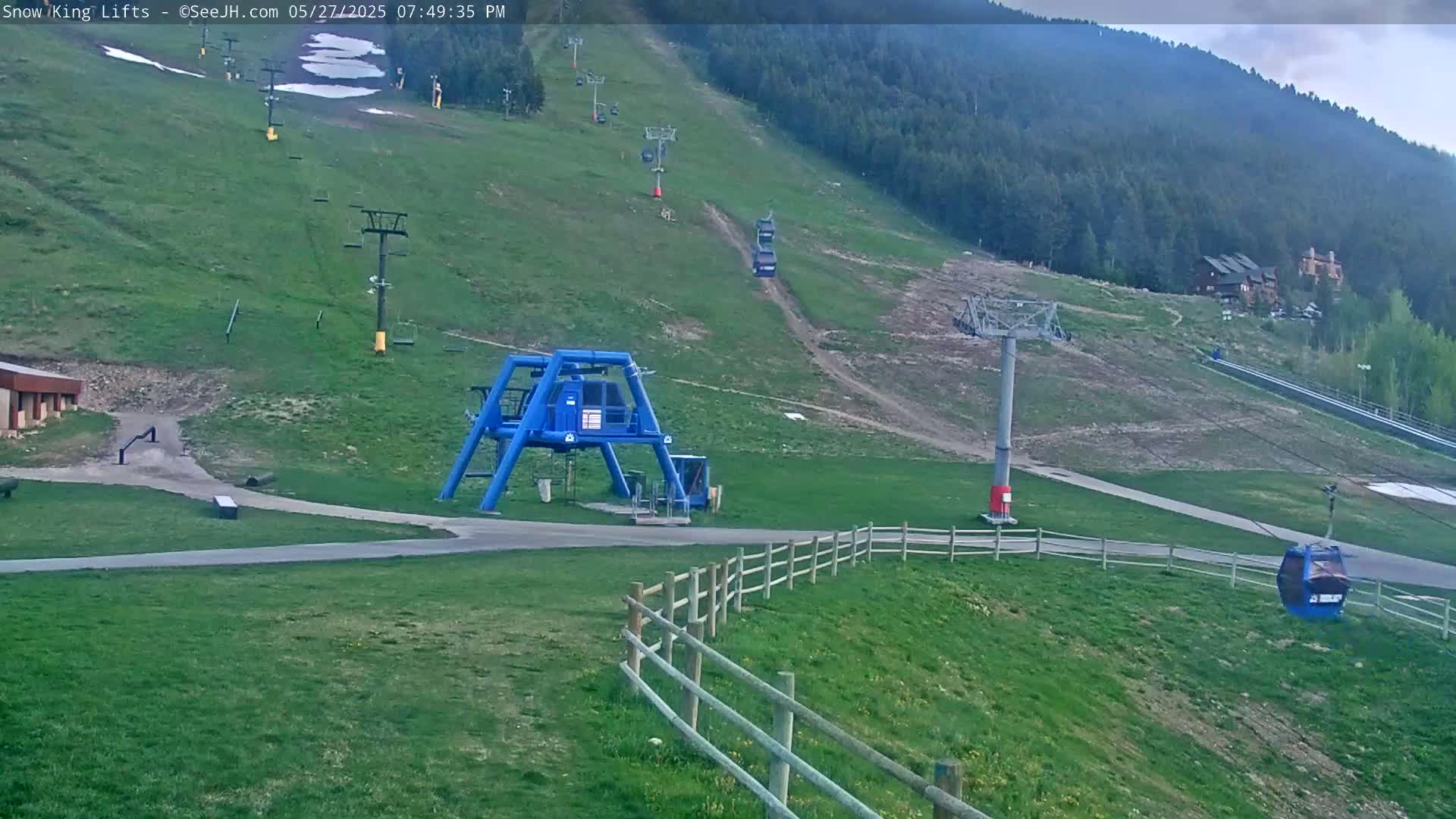 A mostly sunny view of a ski lift station with several chairlifts ascending a grassy mountainside.