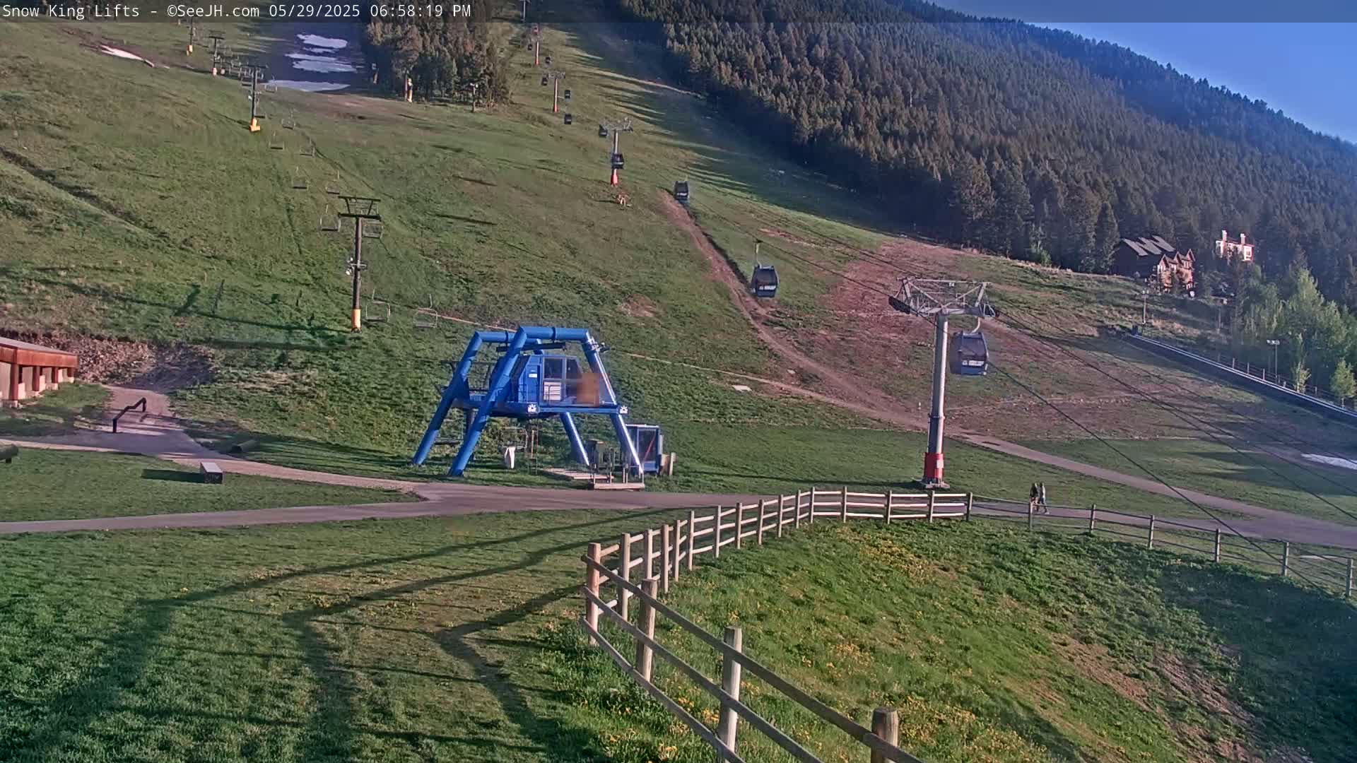 A grassy ski lift area with several chairlifts and a lift station is visible under sunny conditions; a forested mountainside is in the background.