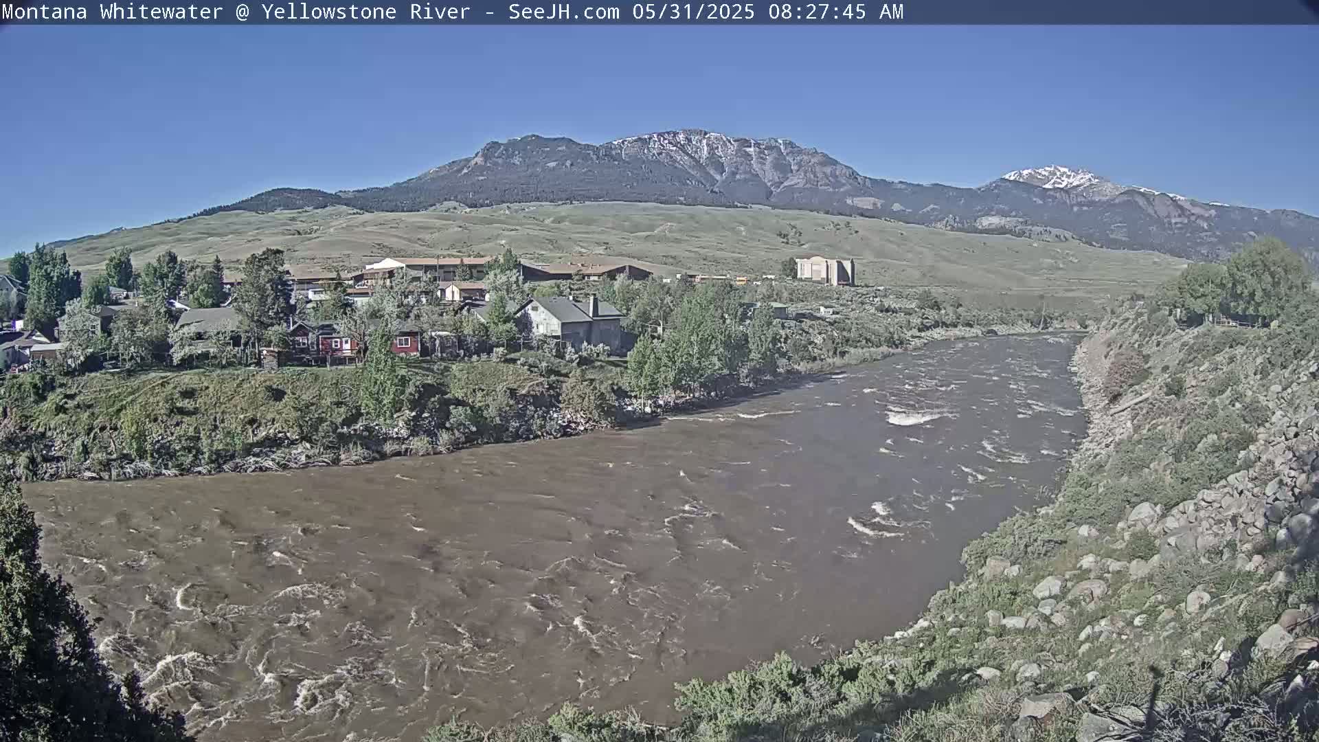 A muddy river flows past a small town nestled at the base of snow-capped mountains under a clear blue sky.
