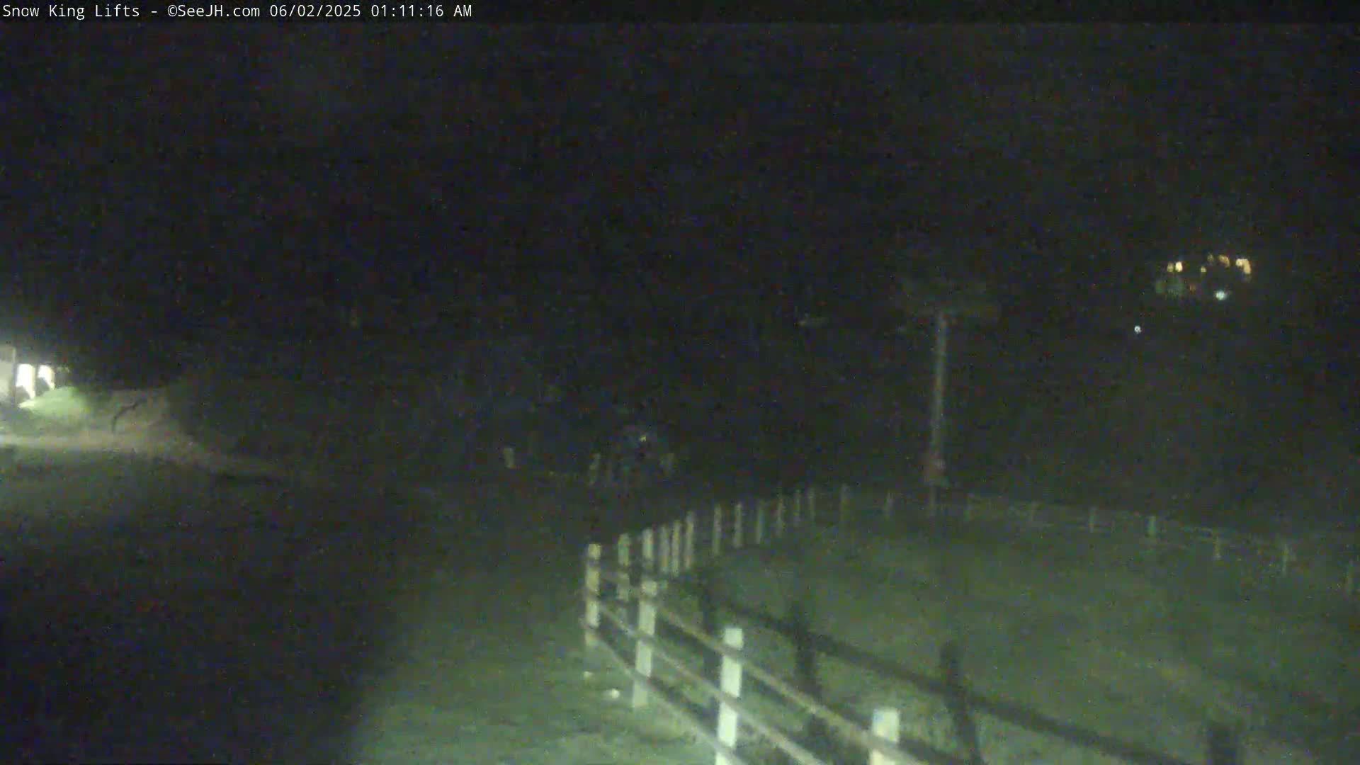 A nighttime, low-light view of a dark area with a white fence, some indistinct objects in the mid-ground, and distant lights.
