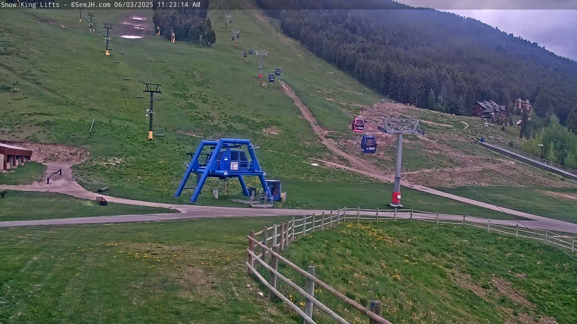 A mostly sunny view of a ski lift base area with several chairlifts ascending a grassy hillside dotted with trees.
