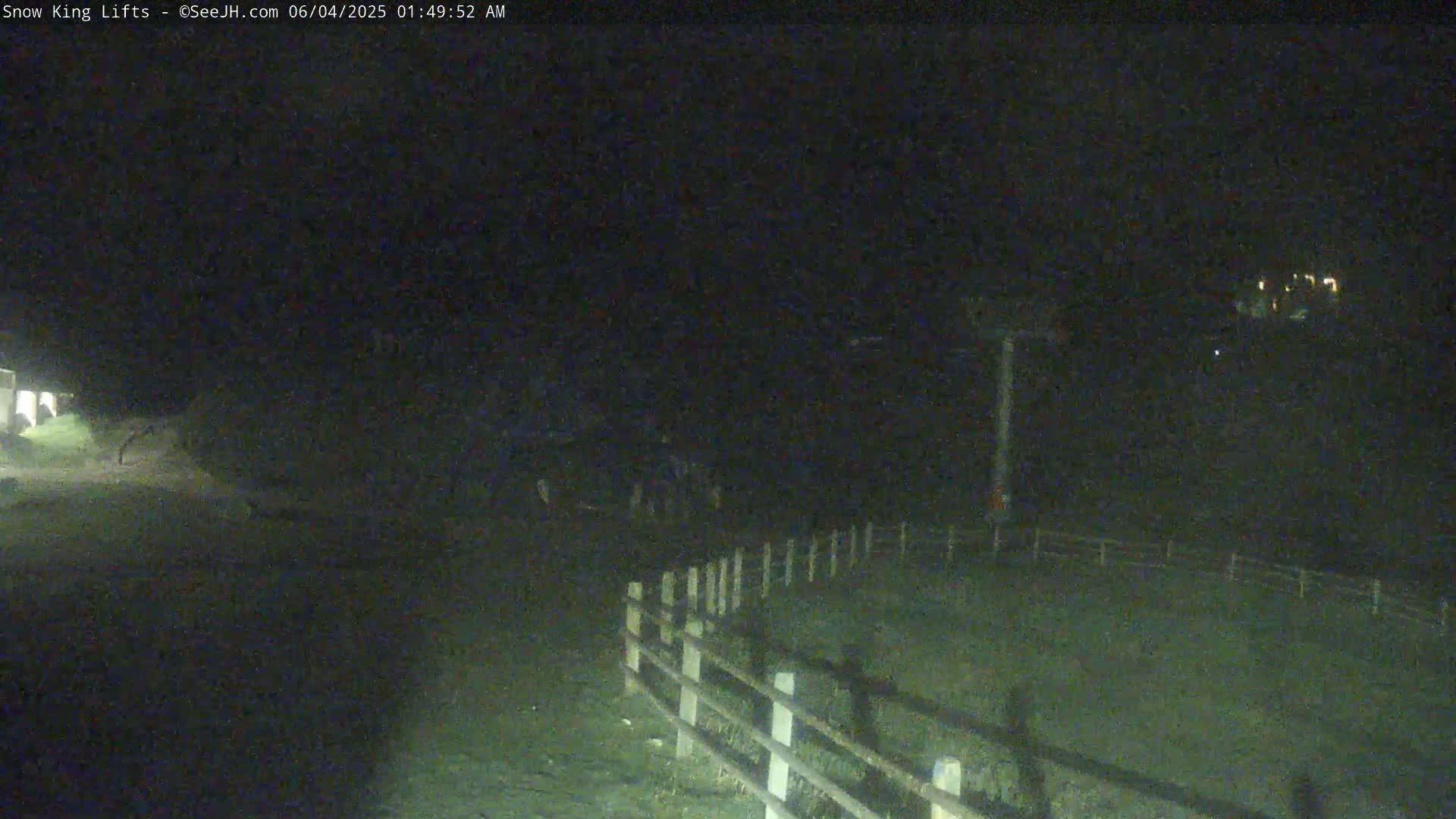 A nighttime, low-light view of a snow-covered area with a fence and a lift visible in the distance.
