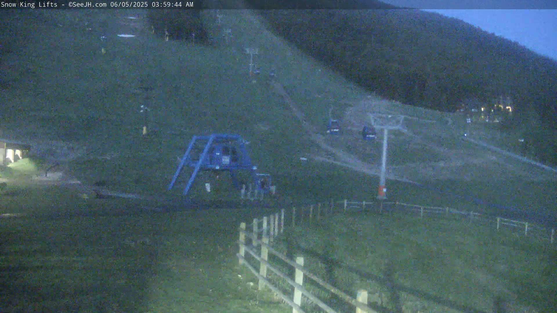 A nighttime view of a ski slope with a chairlift and a few small structures, under a dark sky.