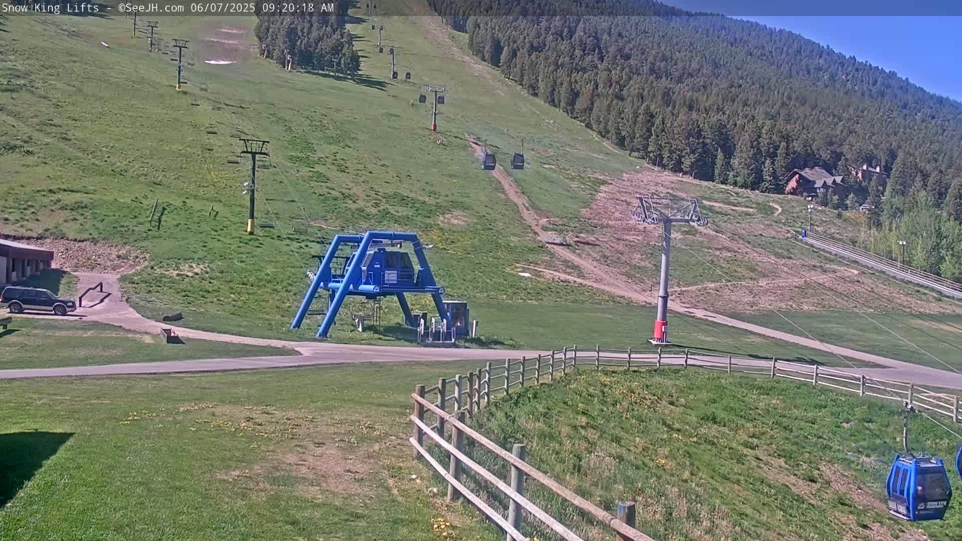 A sunny view of a ski lift area with several chairlifts and a small blue gondola, set against a backdrop of green hills and a distant forest.