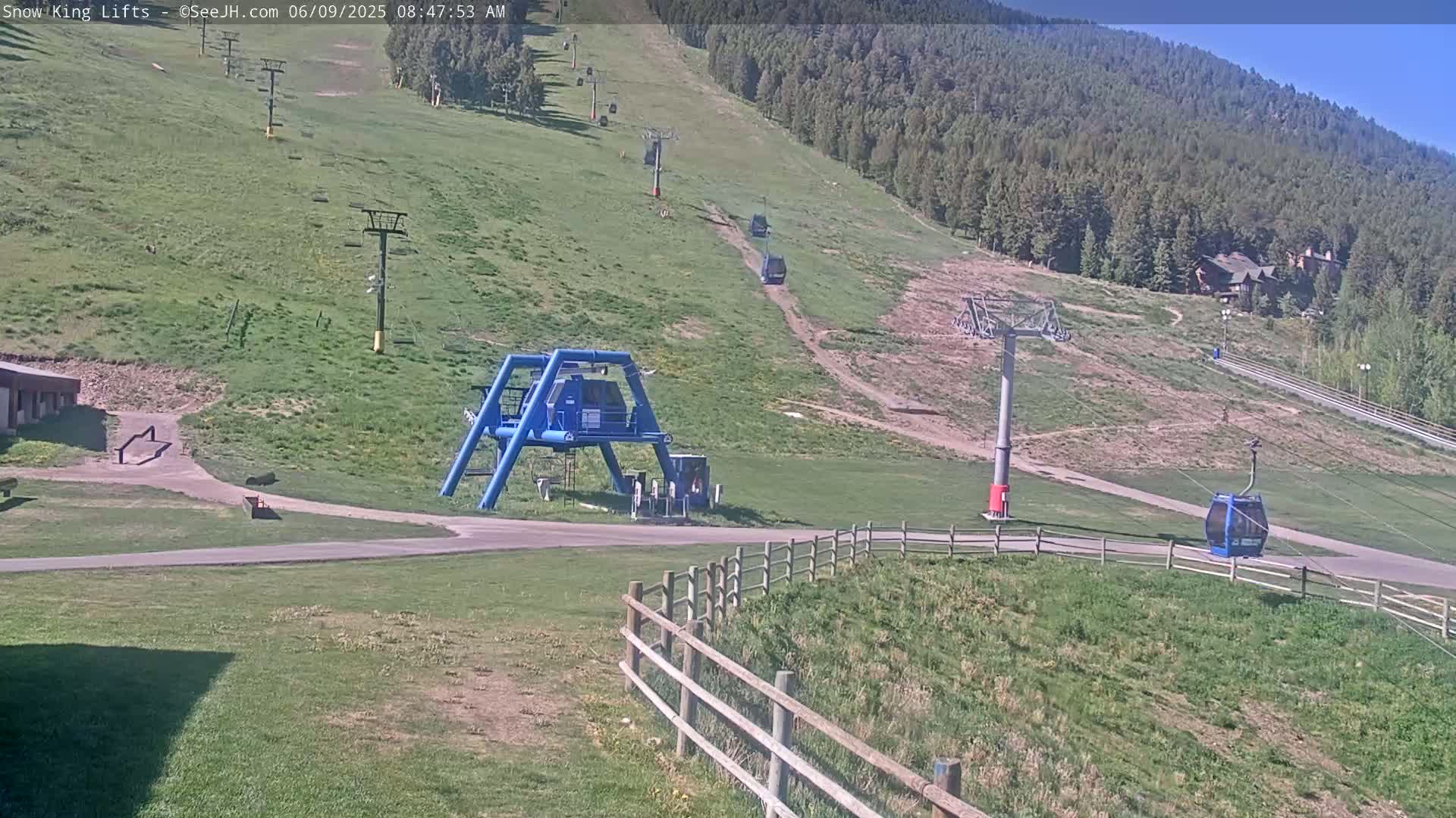 A sunny view of a ski lift area with grassy hills, ski lifts, and a chairlift, in the foreground is a blue ski lift terminal.