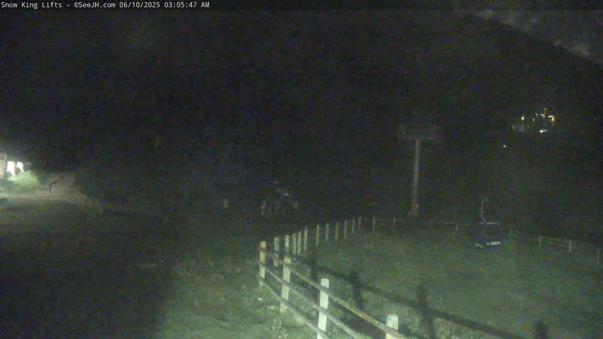 A dark, nighttime scene shows a grassy area enclosed by a wooden fence, with a vehicle and indistinct shapes in the distance under a dark sky.