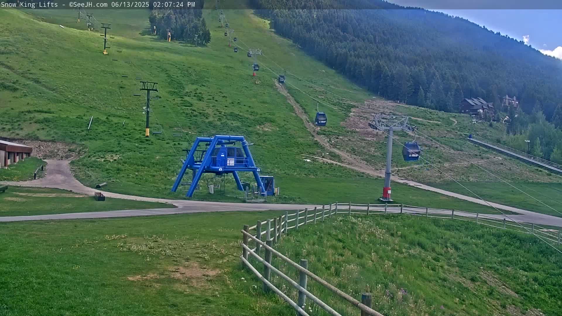 A mostly sunny view of a ski lift base area with several chairlifts ascending a grassy hillside into a forest.