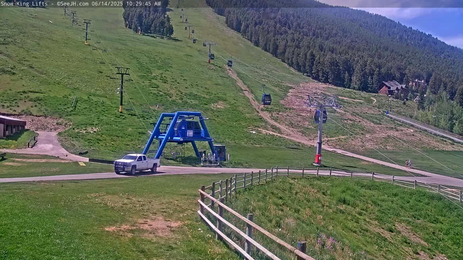 A white truck is parked near a ski lift base at the bottom of a grassy hill dotted with several chairlifts ascending a mountainside under a partly sunny sky.