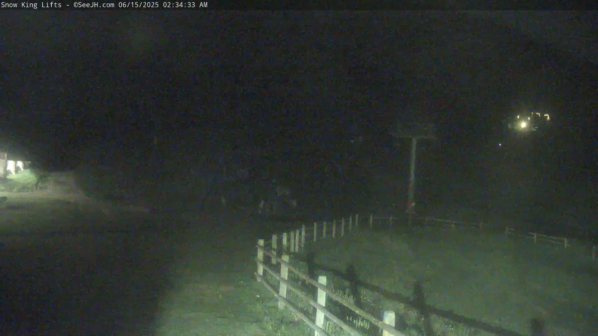 A nighttime, low-light image shows a dark, grassy area with a short, white-painted fence, and a distant, dimly lit structure.