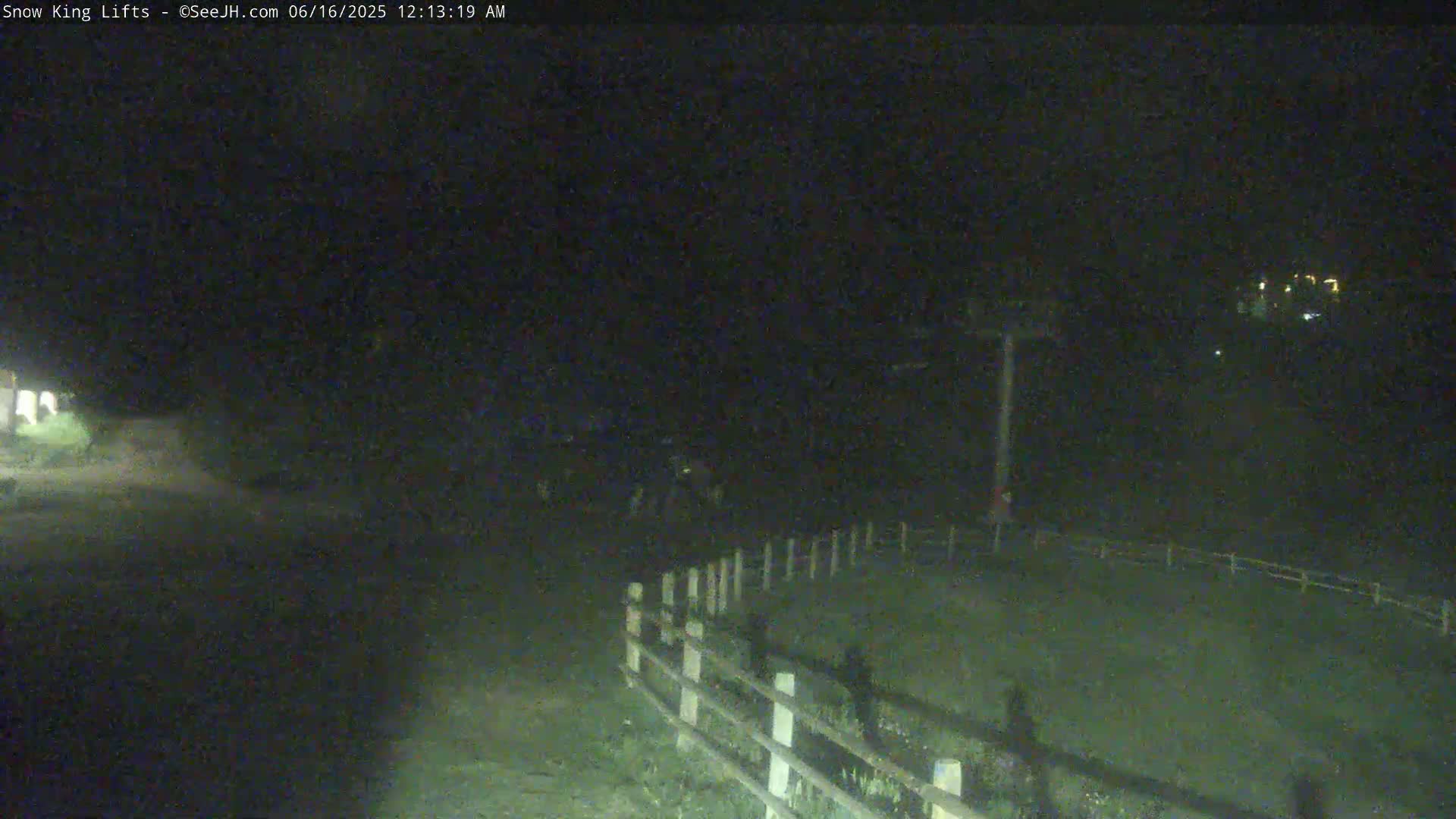 A nighttime, low-light view of a mostly dark area with a wooden fence, a tall structure in the distance that appears to be a ski lift, and some dimly lit areas suggesting buildings or lights further away.