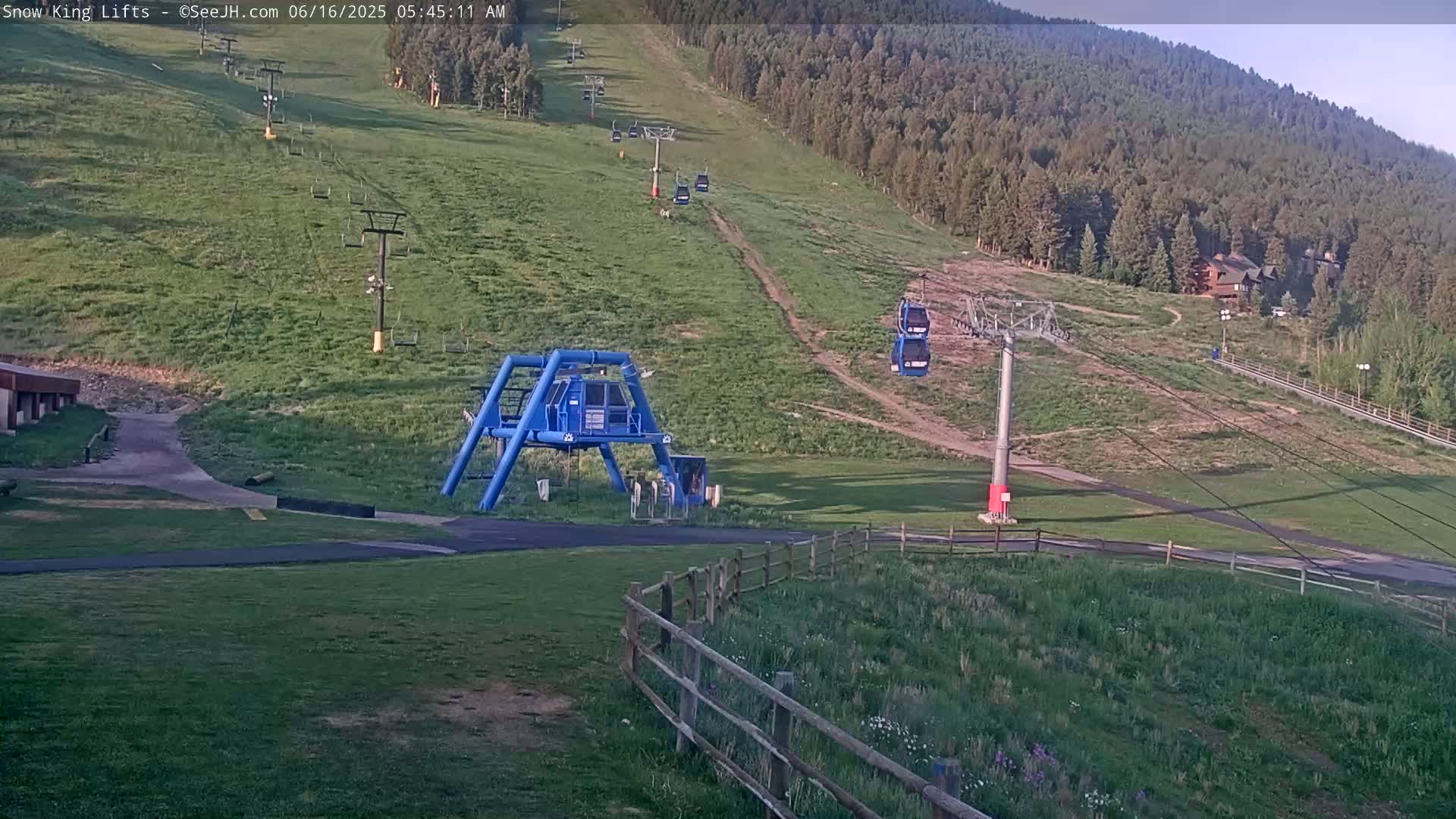 A ski lift base station and several chairlifts are situated on a grassy hillside under a clear sky, with a forested mountain in the background.