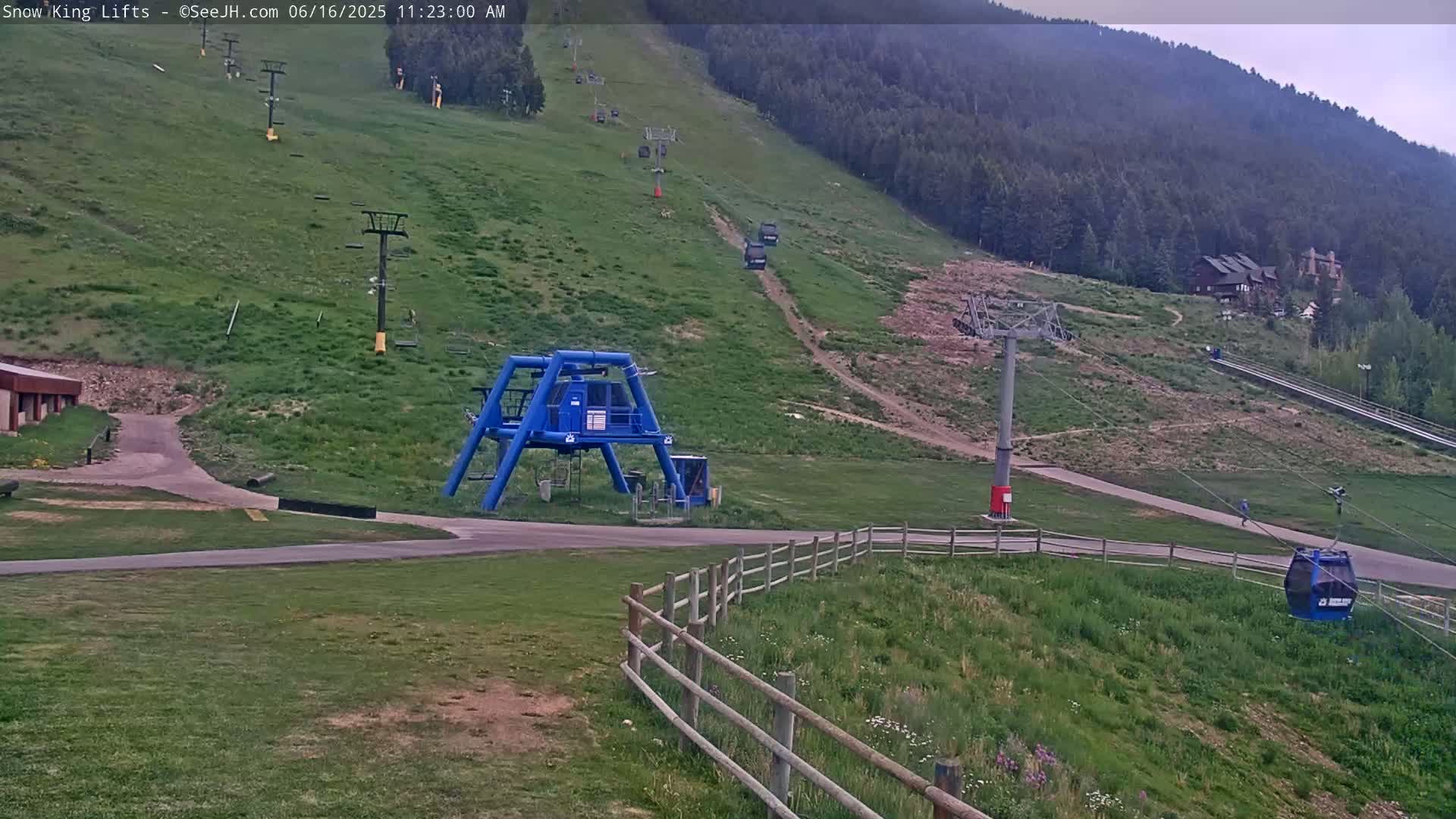 A mostly clear, summer day reveals a ski lift base area with several chairlifts ascending a grassy mountainside, along with a blue lift maintenance structure and a gondola car.