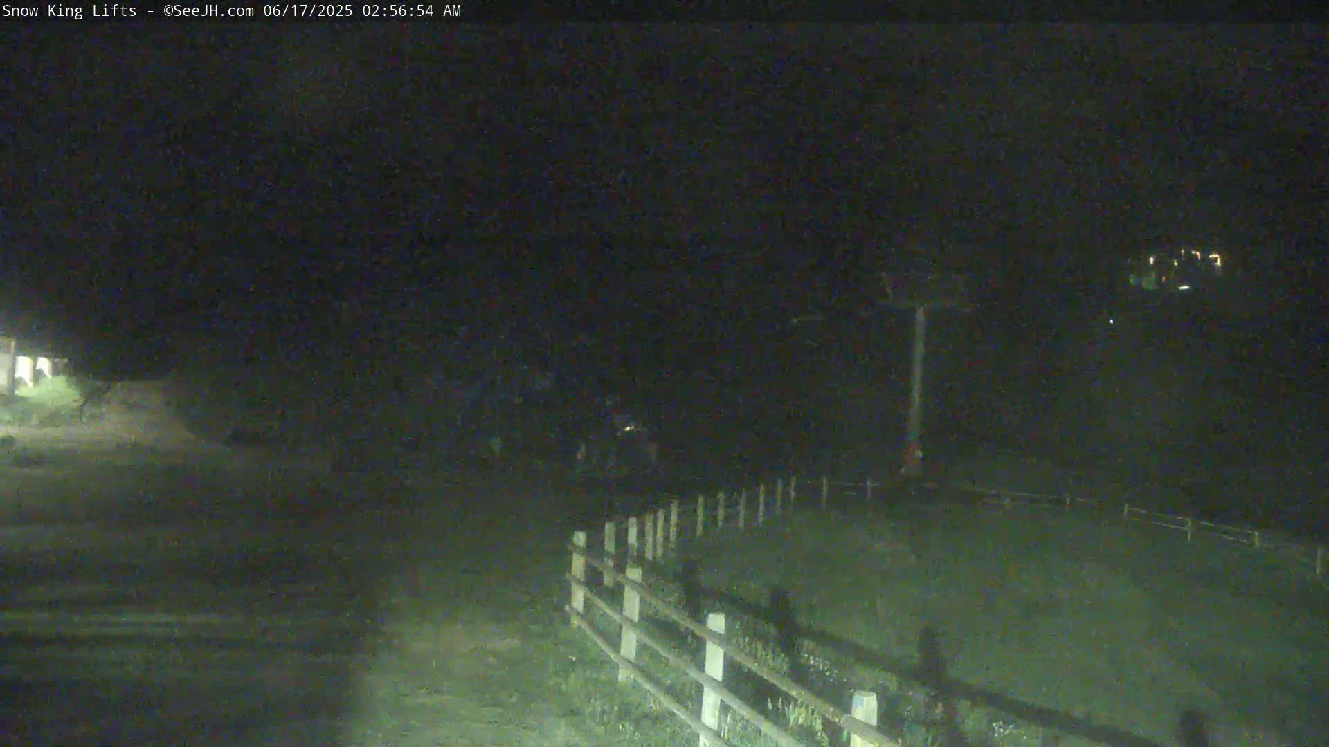 A nighttime, low-light image shows a grassy area enclosed by a wooden fence, with a tall pole-like structure visible in the middle ground and faint lights in the distance.