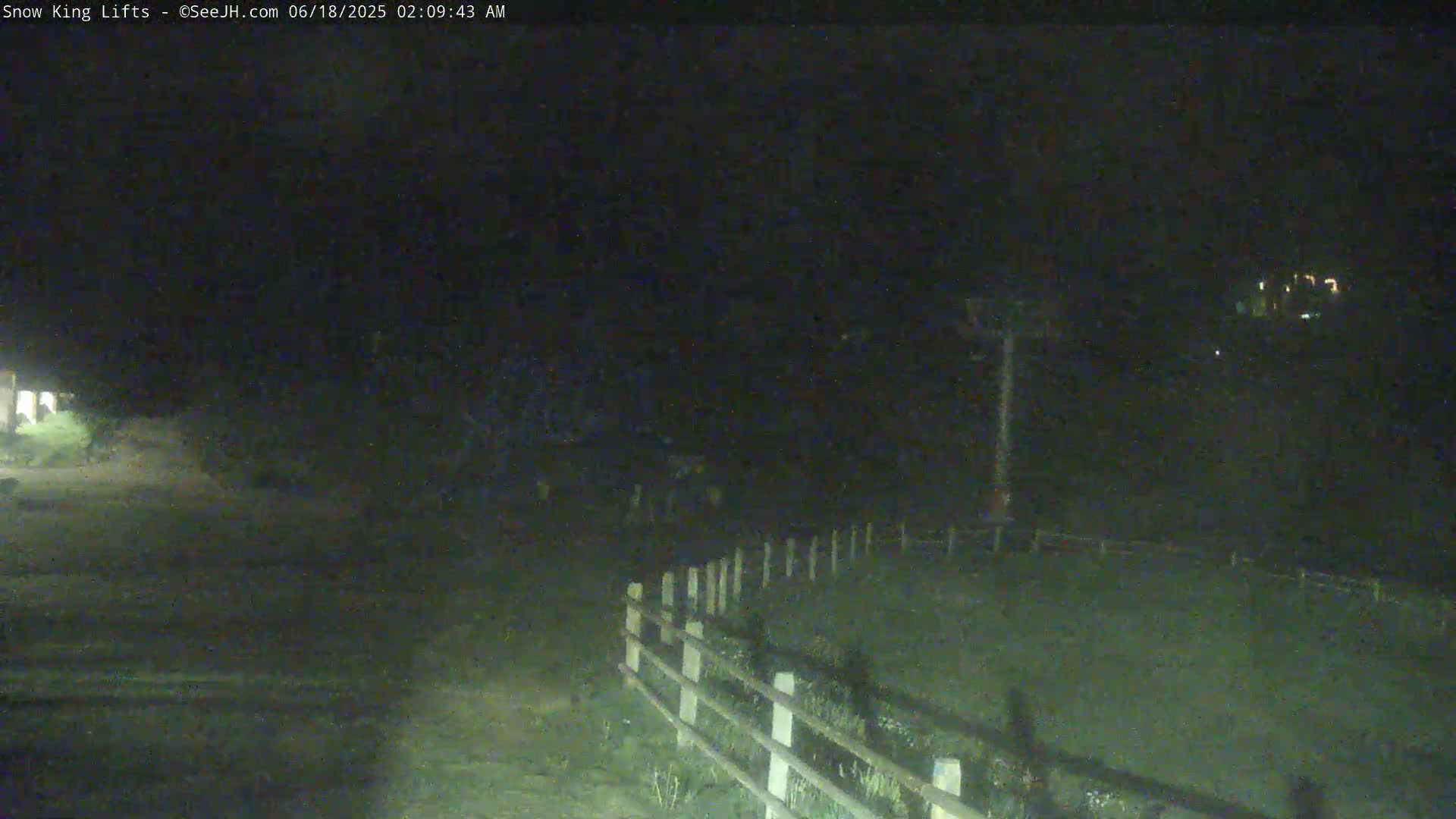 A dark, nighttime scene shows a sparsely lit area with a short, white fence and a ski lift in the distance.