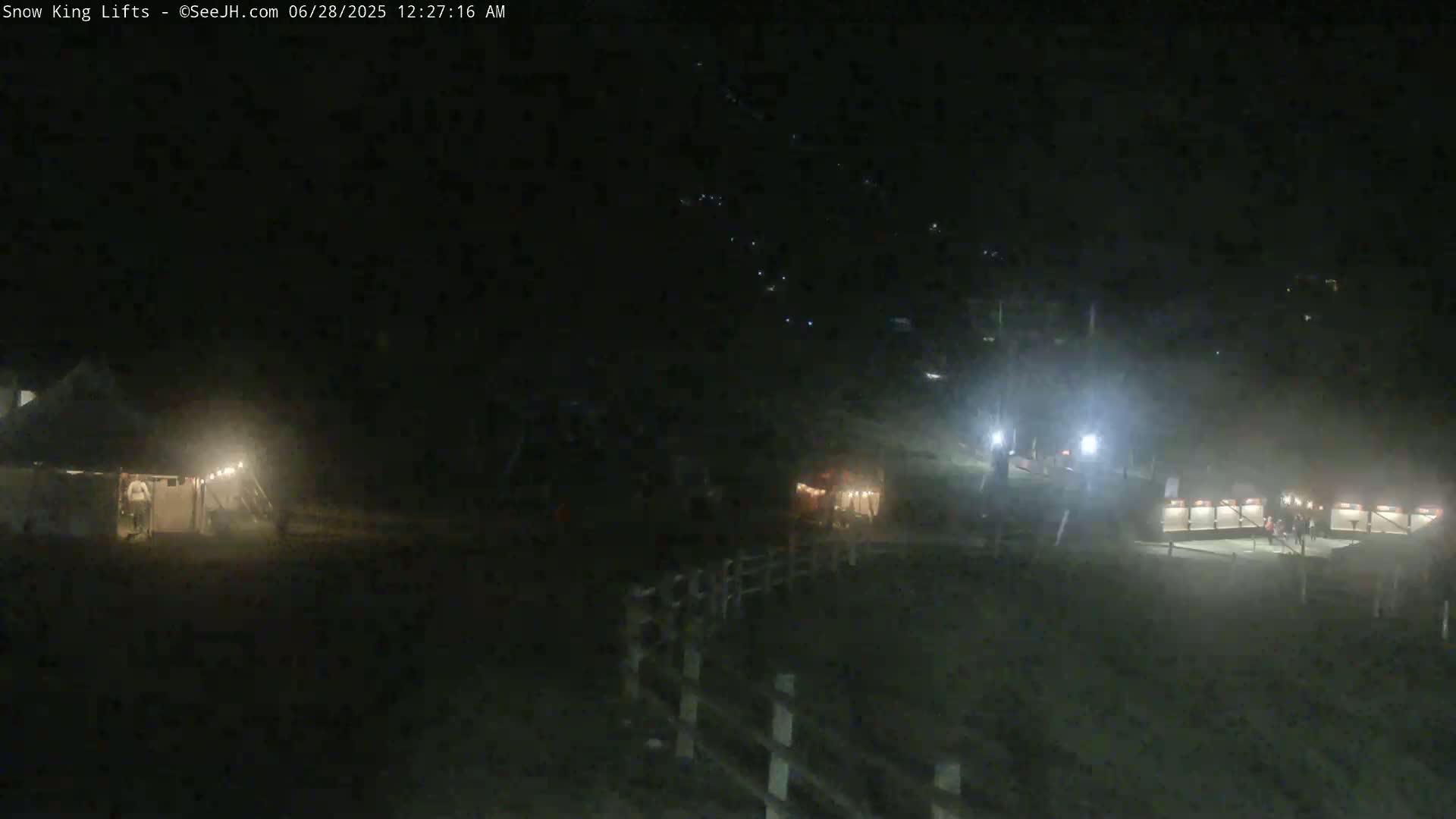 At night, dimly lit structures and a few people are visible in a hazy, dark outdoor scene.