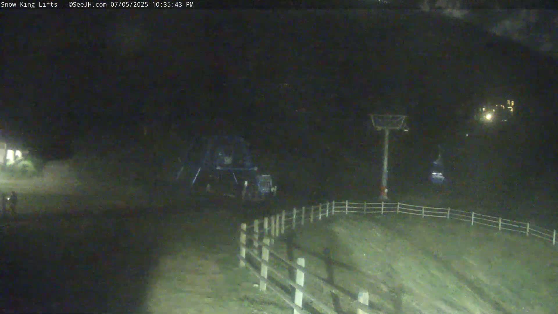 A nighttime view of a ski lift and surrounding area, possibly showing a chairlift in operation with a dark, possibly overcast sky.
