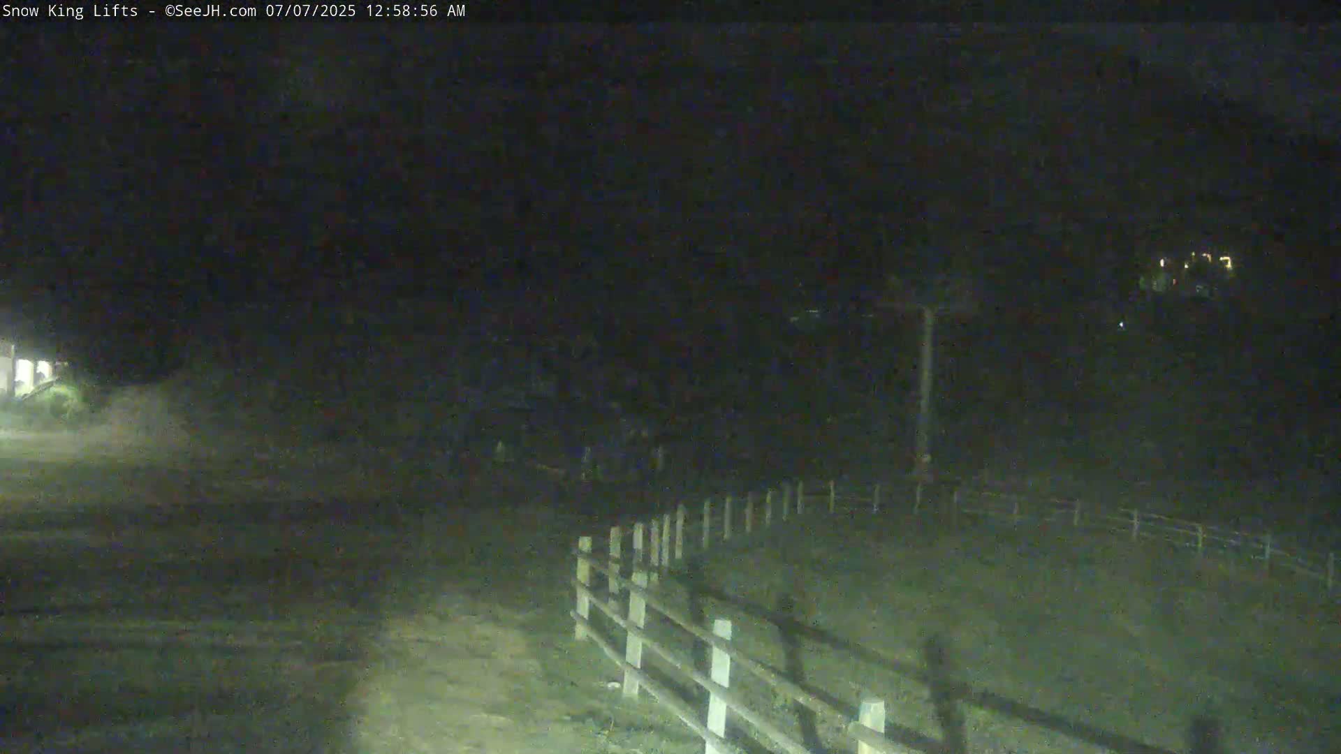 A nighttime, low-light view shows a dirt road next to a wooden fence, with dark, indistinct shapes in the background under a dark sky.