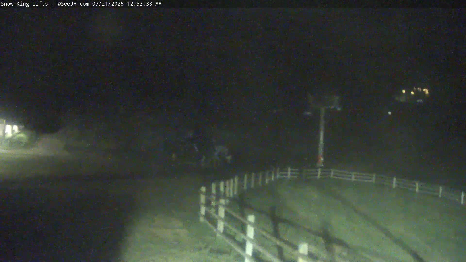 A nighttime, low-light view shows a snow-covered slope with a ski lift tower and a fence in the foreground.