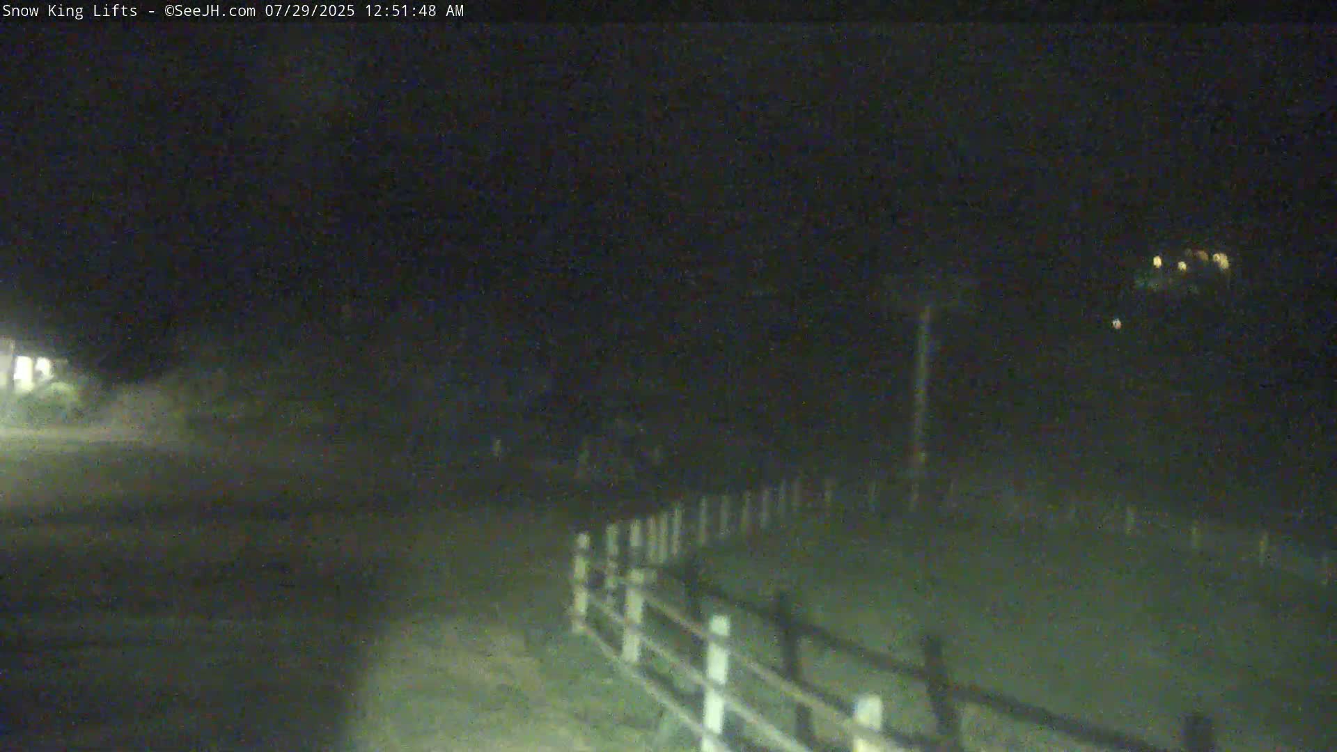 A nighttime, low-light image shows a dark road and a white fence, with some distant lights visible in the background.