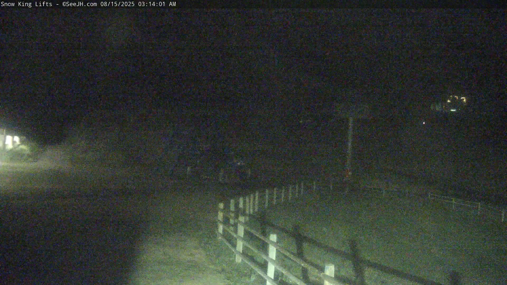 A nighttime, low-light image shows a dark road and a white fence, with some distant lights visible in the background.