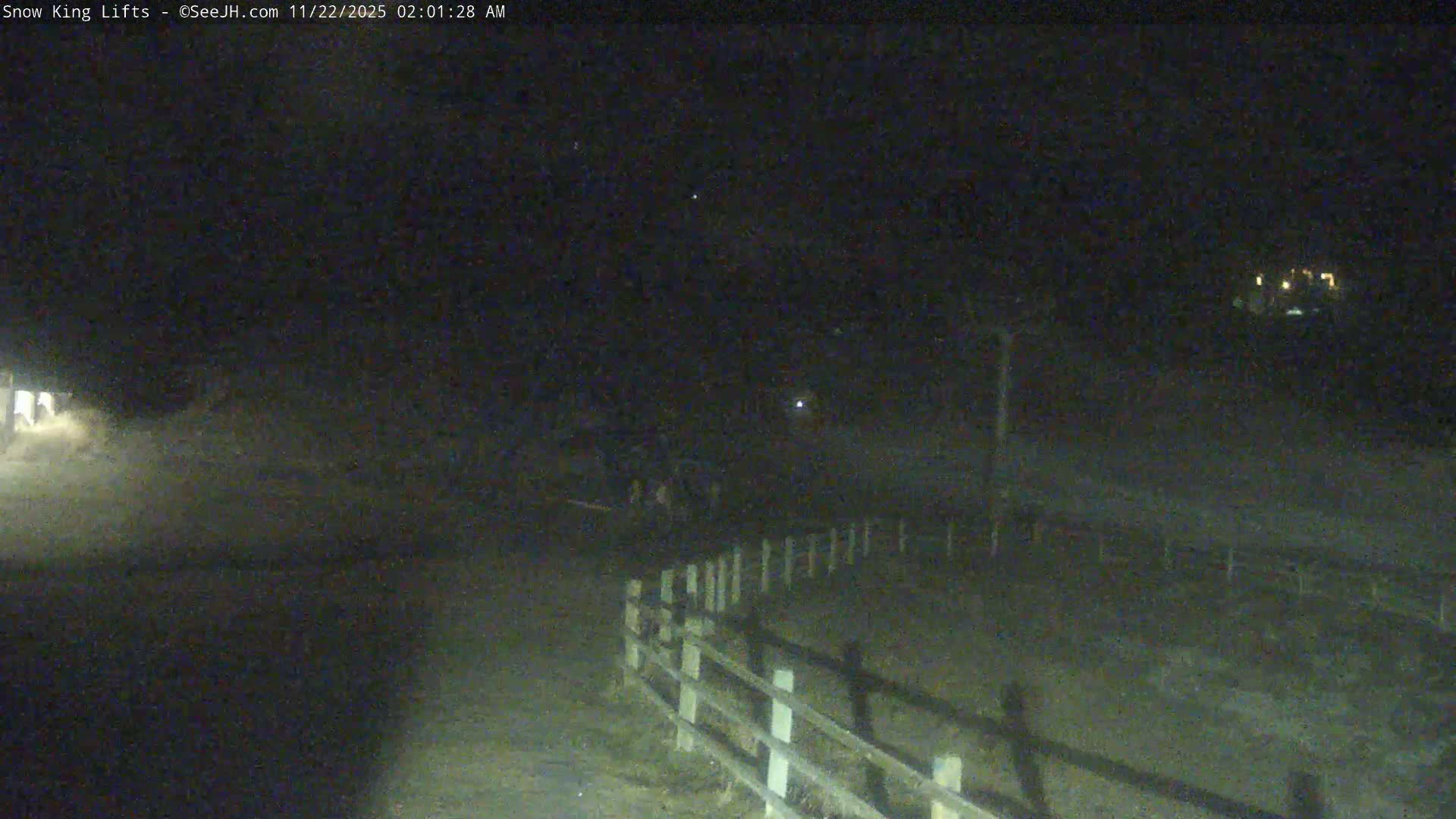A grainy, dark nighttime view shows a wooden fence curving into the middle ground, with faint structures and lights visible in the distance under clear skies.