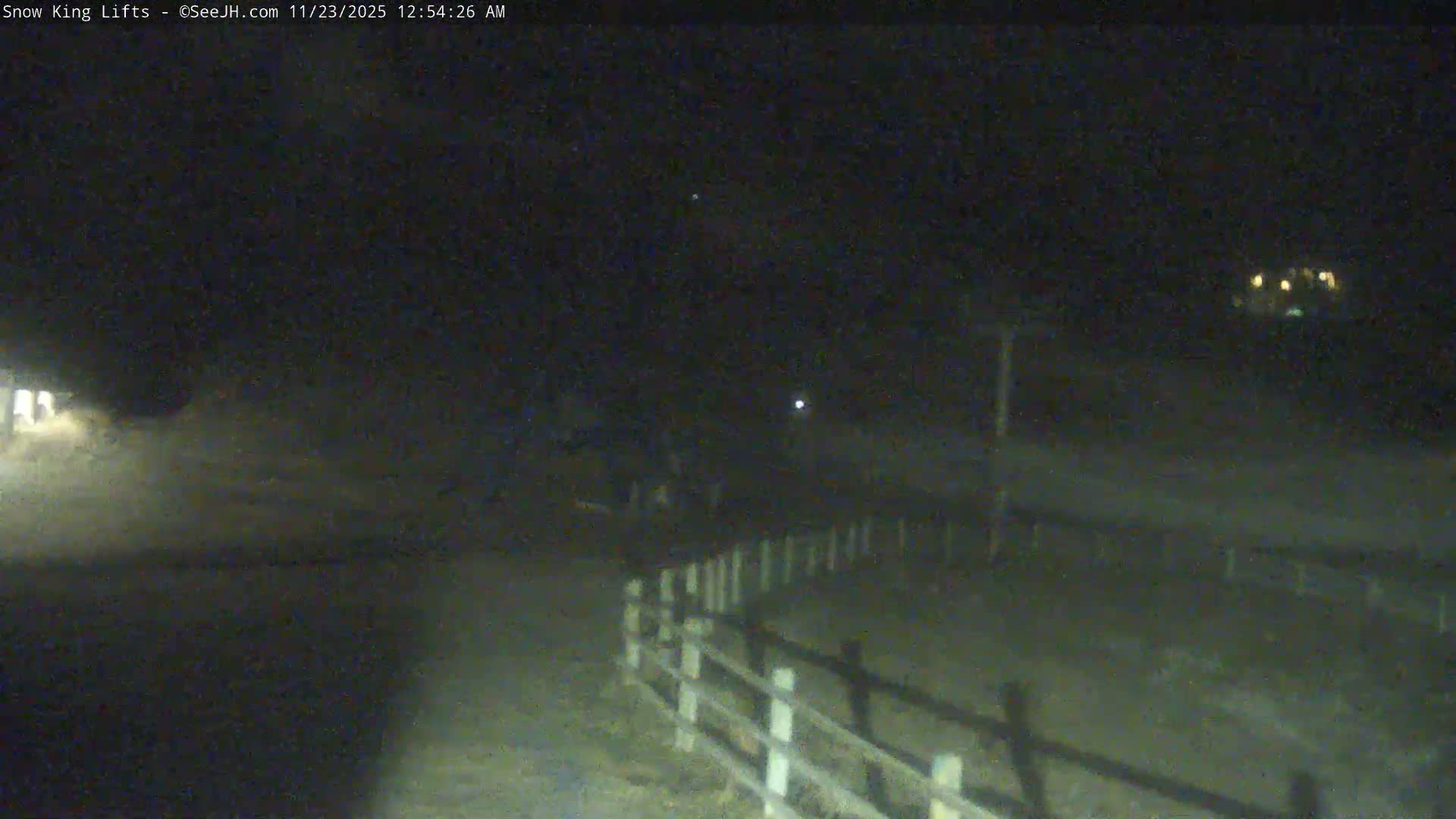 A dim, grainy nighttime view of an outdoor area features a wooden fence in the foreground, illuminated patches of ground, and scattered lights from distant structures on a clear night.