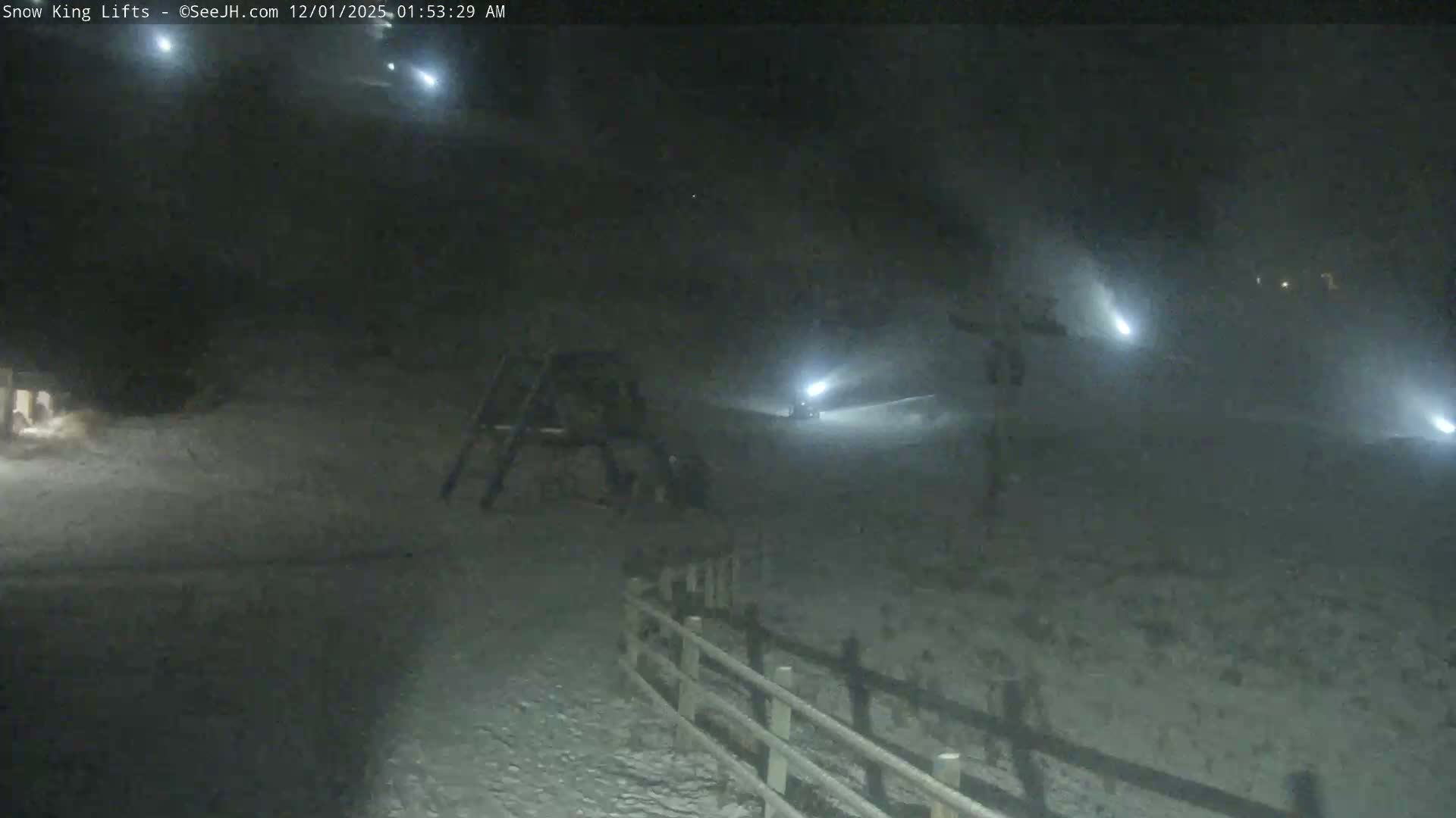 A dark, snowy ski area is illuminated at night by several bright snowmaking lights producing visible plumes of mist, with a wooden fence in the foreground, suggesting cold, active snowmaking conditions.