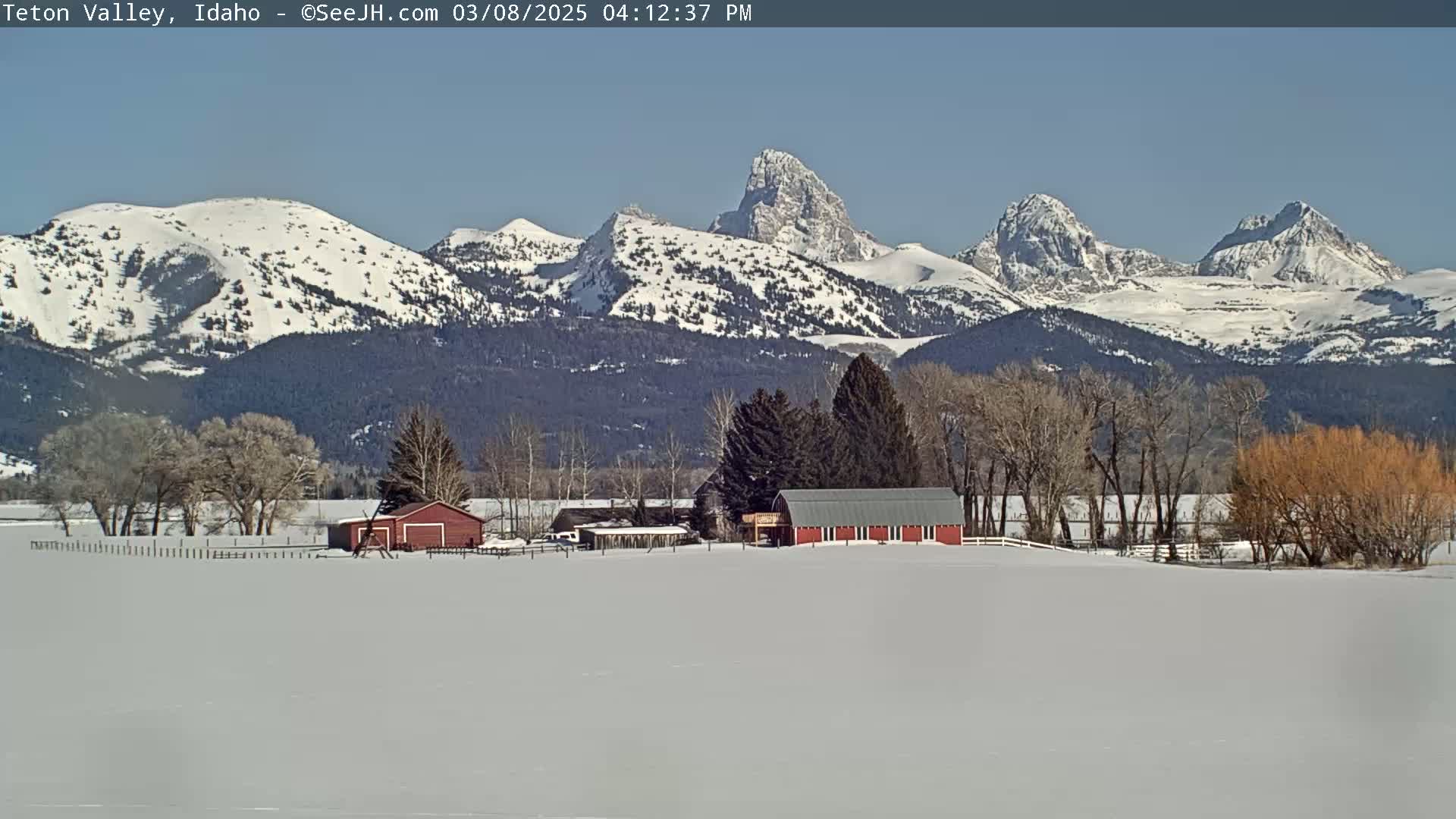 A snow-covered landscape features a red barn and other farm buildings in the foreground, with snow-capped mountains rising in the background under a clear, sunny sky.